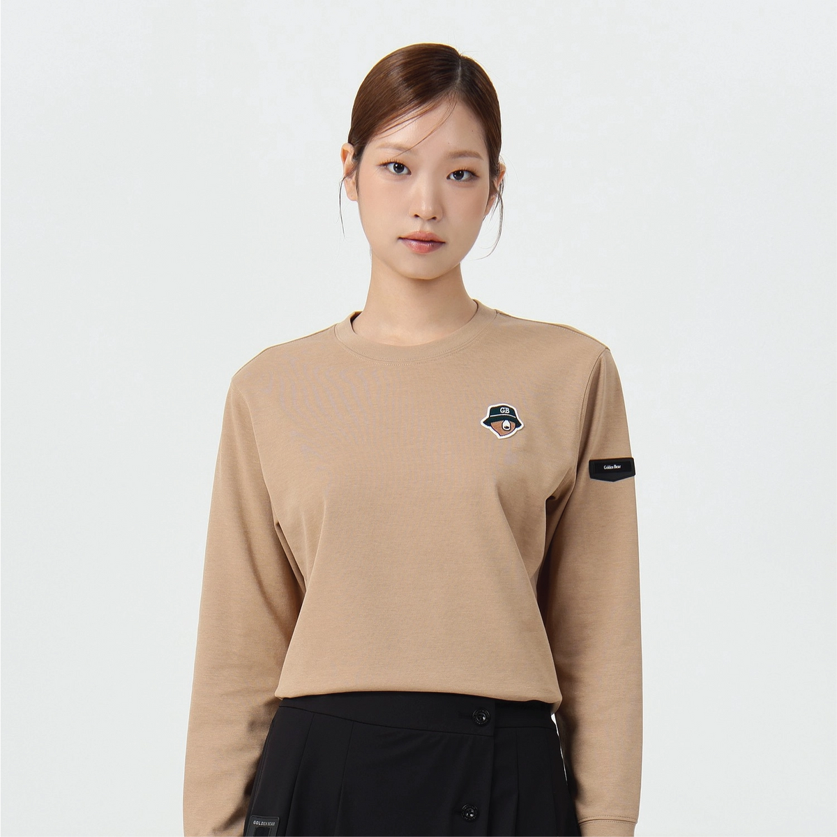 A woman modeling a beige long-sleeve crewneck sweatshirt with a small embroidered logo on the left chest and a black sleeve patch, paired with a black skirt against a plain light gray background.