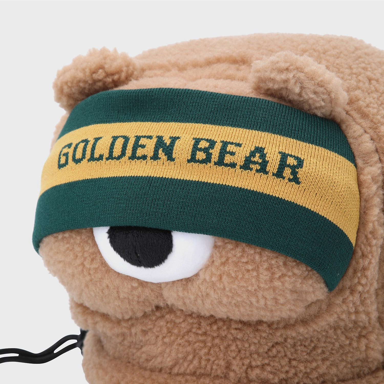 Close-up of a plush teddy bear head with a knit headband across the eyes. The headband is dark green with a wide yellow stripe and the words 'GOLDEN BEAR' in dark green lettering.