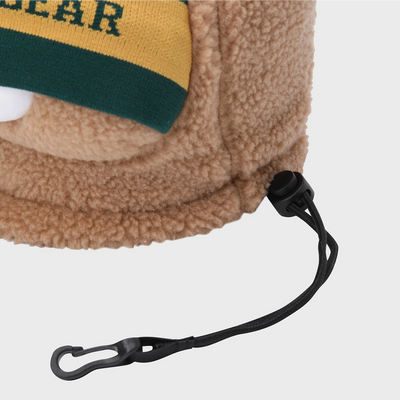 Close-up of a tan sherpa fleece hat with a yellow and green knit cuff and a black drawstring carabiner, shown on a light gray background.