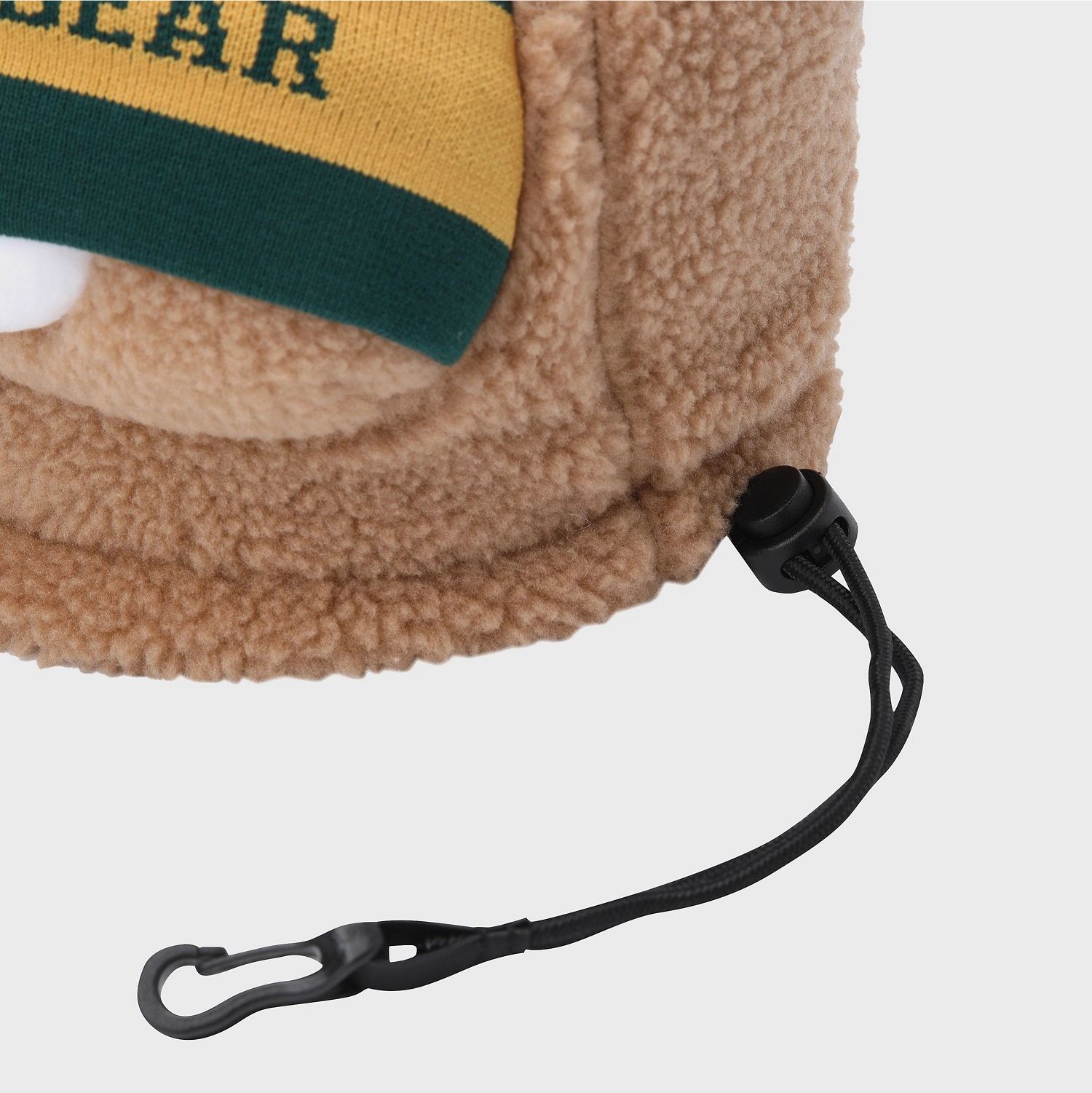 Close-up of a tan sherpa fleece hat with a yellow and green knit cuff and a black drawstring carabiner, shown on a light gray background.