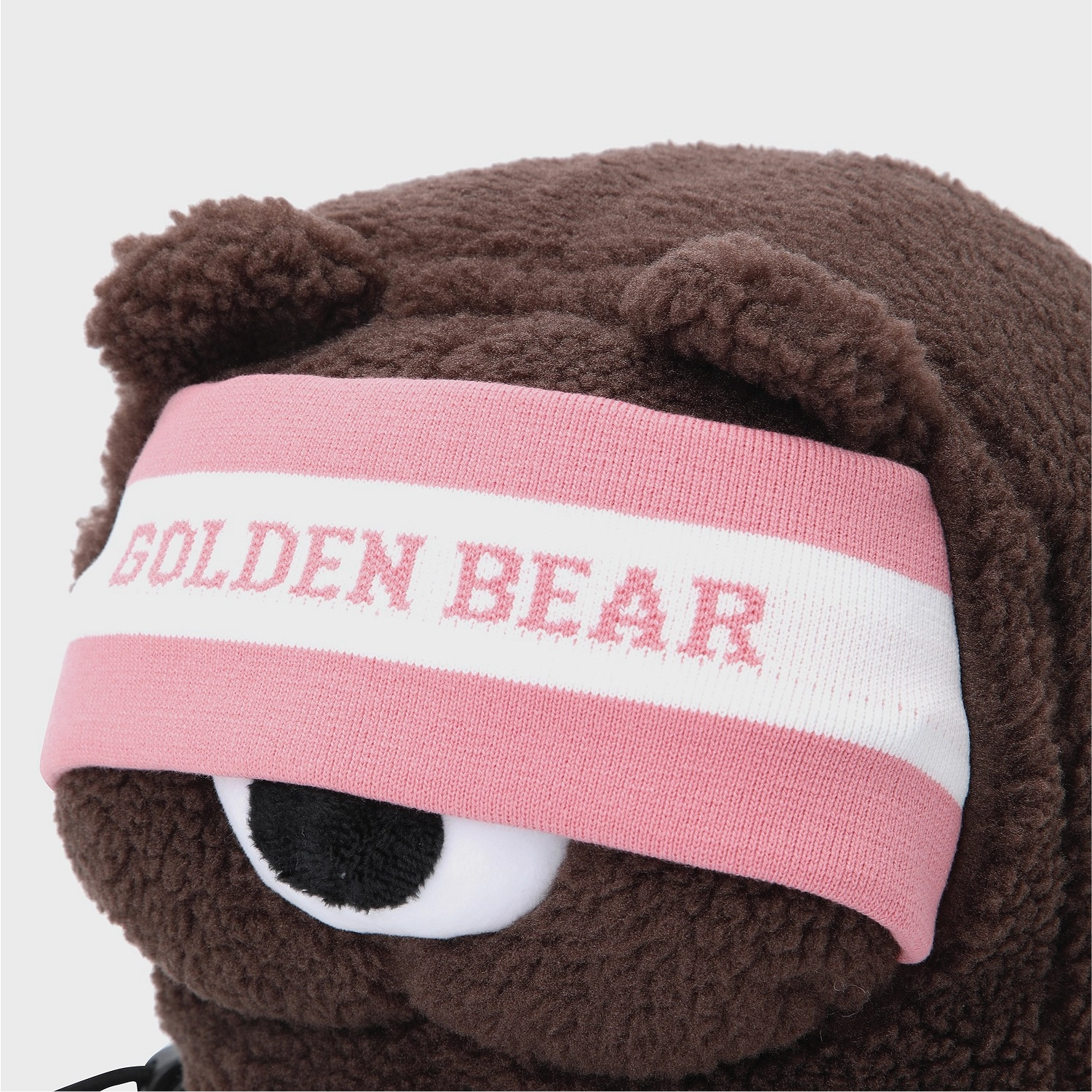 Close-up of a brown plush bear wearing a pink and white knit headband with the words 'GOLDEN BEAR' in pink, on a light gray background.