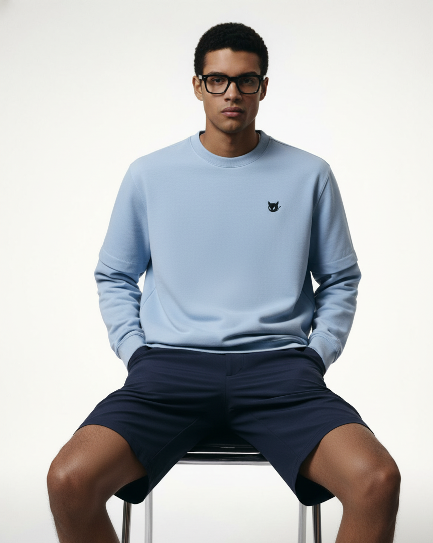 Man wearing a light blue sweatshirt and navy shorts sitting on a white stool against a white background