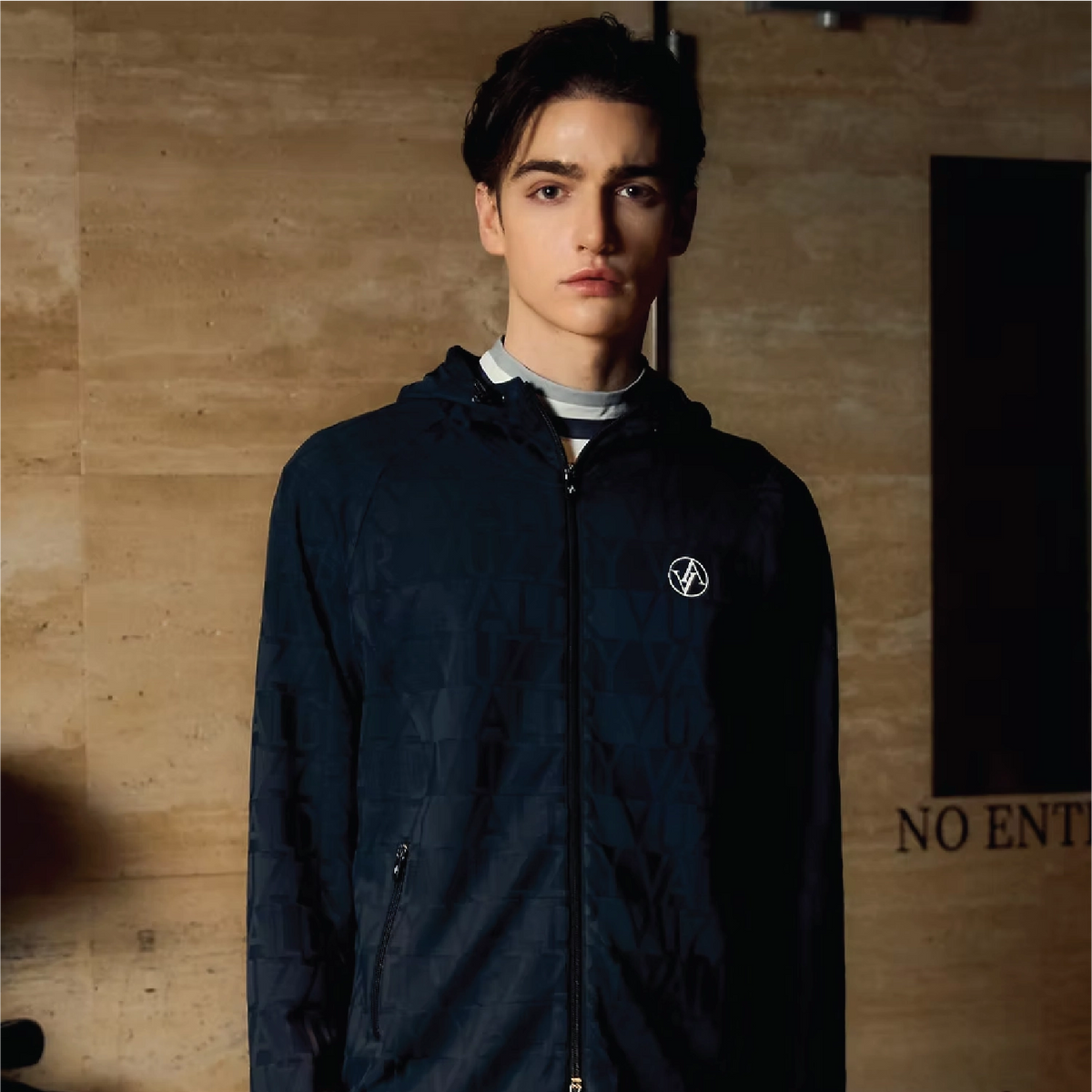 A man wearing a dark navy quilted zip-up jacket with a white circular logo on the chest, layered over a white turtleneck, standing against a textured beige wall.