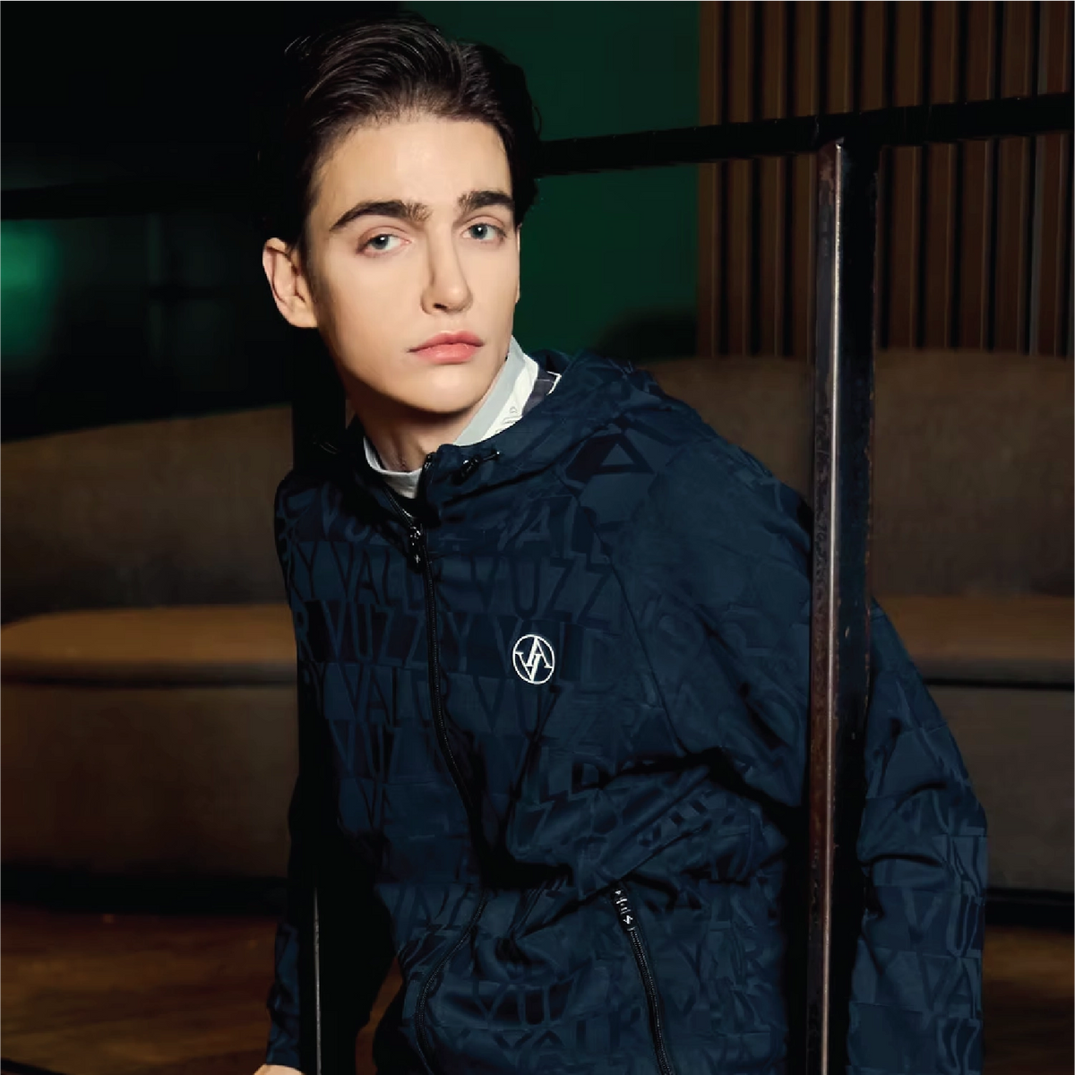 A man wearing a dark navy blue patterned hooded jacket with a white circular logo on the chest, posed in a dimly lit indoor setting.