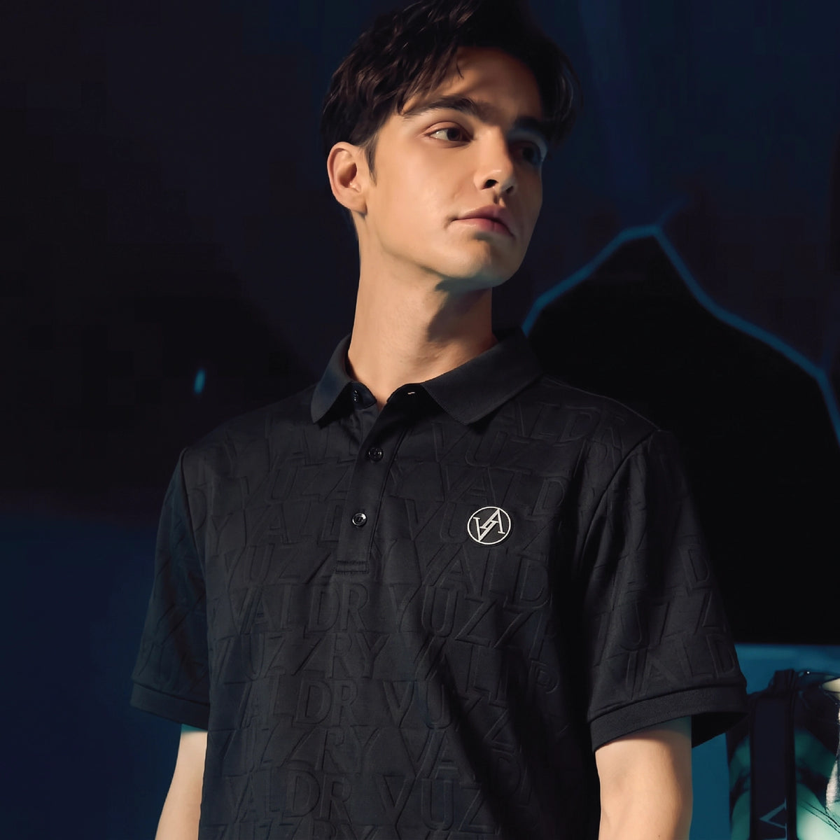 A man wearing a black short-sleeve polo shirt with a subtle tonal geometric pattern and a small circular logo on the left chest, photographed against a dark blue-toned background.