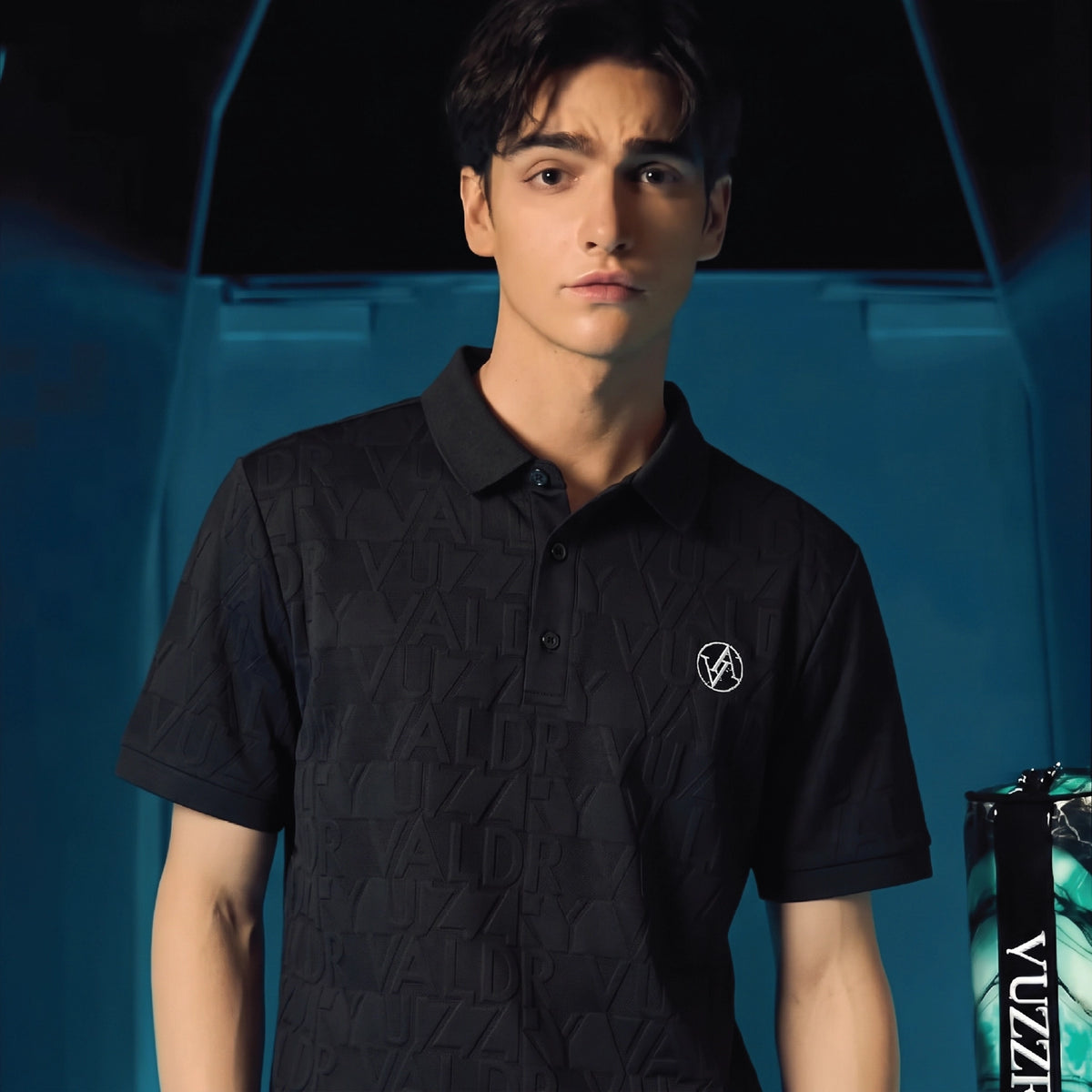 A young man wearing a black short-sleeve polo shirt with a subtle geometric texture and a small white circular logo on the left chest, posed against a dark blue and black background.