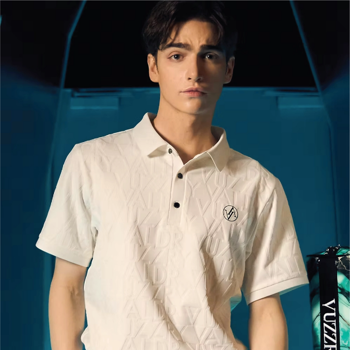 A man wearing a textured off-white short-sleeve polo shirt with a small embroidered logo on the left chest, set against a dark blue and black background.