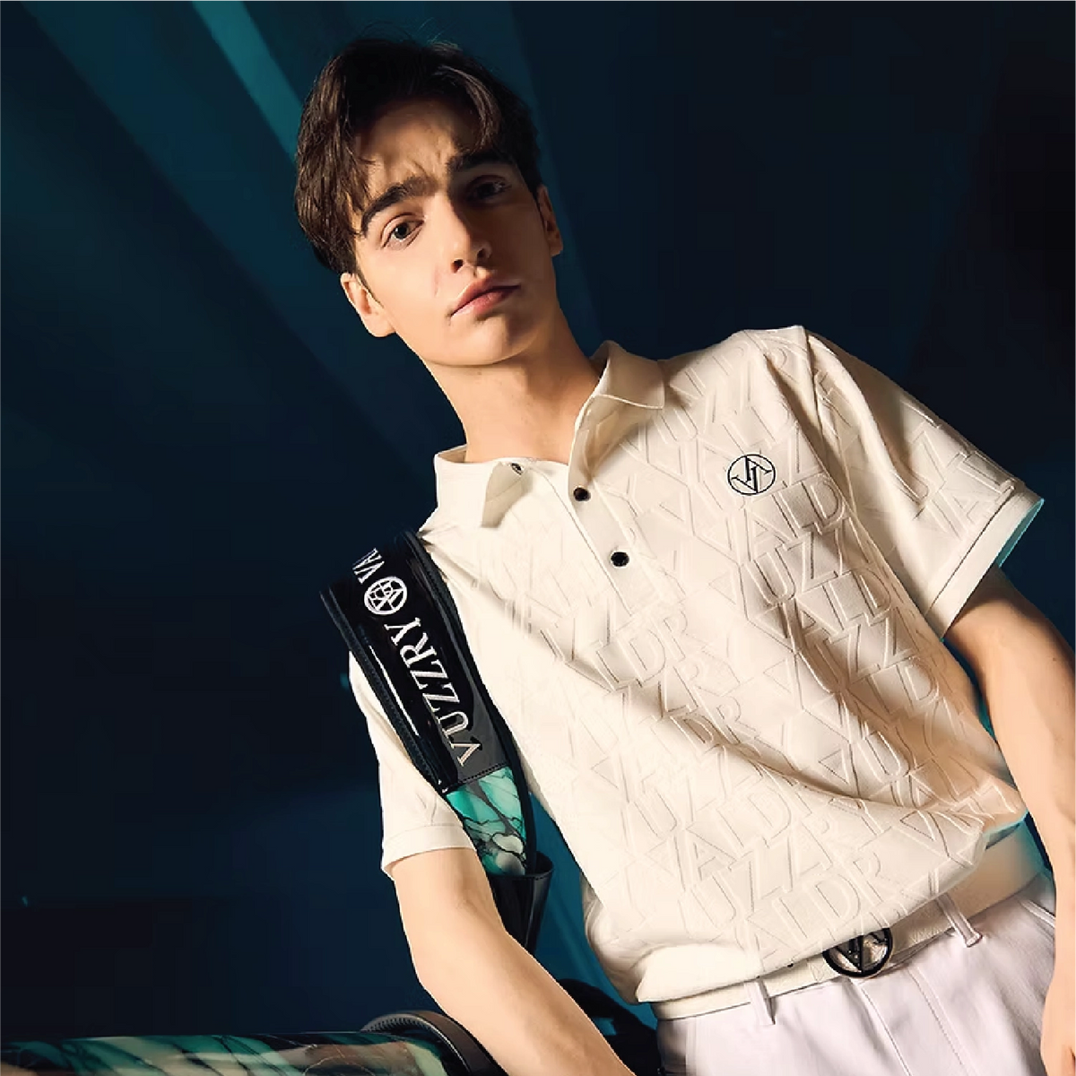 A young man wearing a textured off-white short-sleeve polo shirt with a black and white logo on the chest, paired with white trousers and carrying a black and green bag, set against a dark blue background.