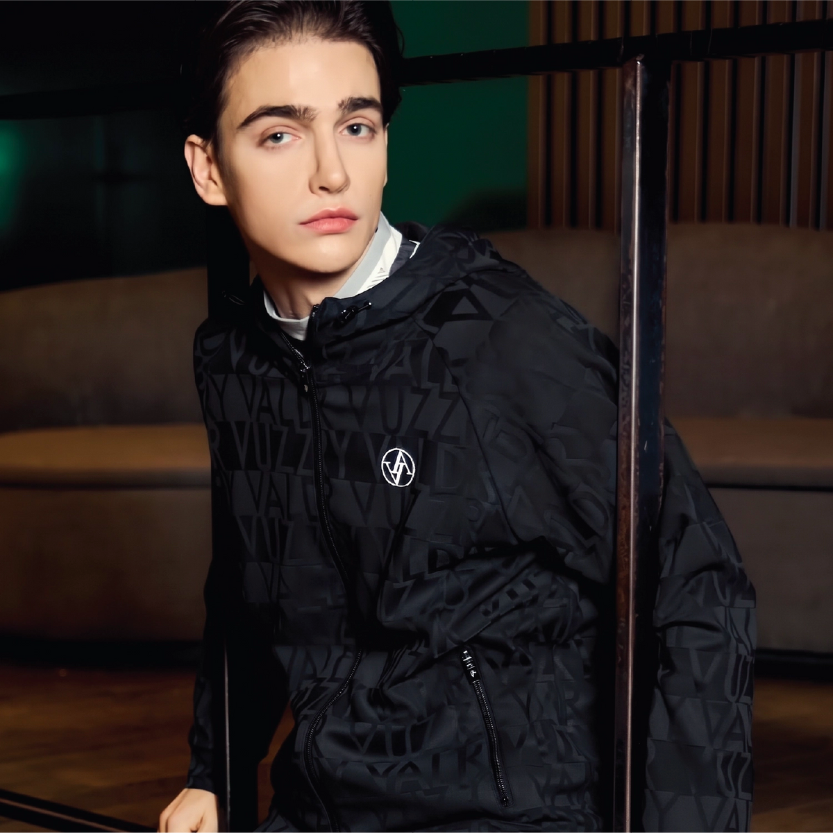 A man wearing a black patterned hooded jacket with a white collar and a small circular logo patch on the chest, posed in a dimly lit indoor setting.