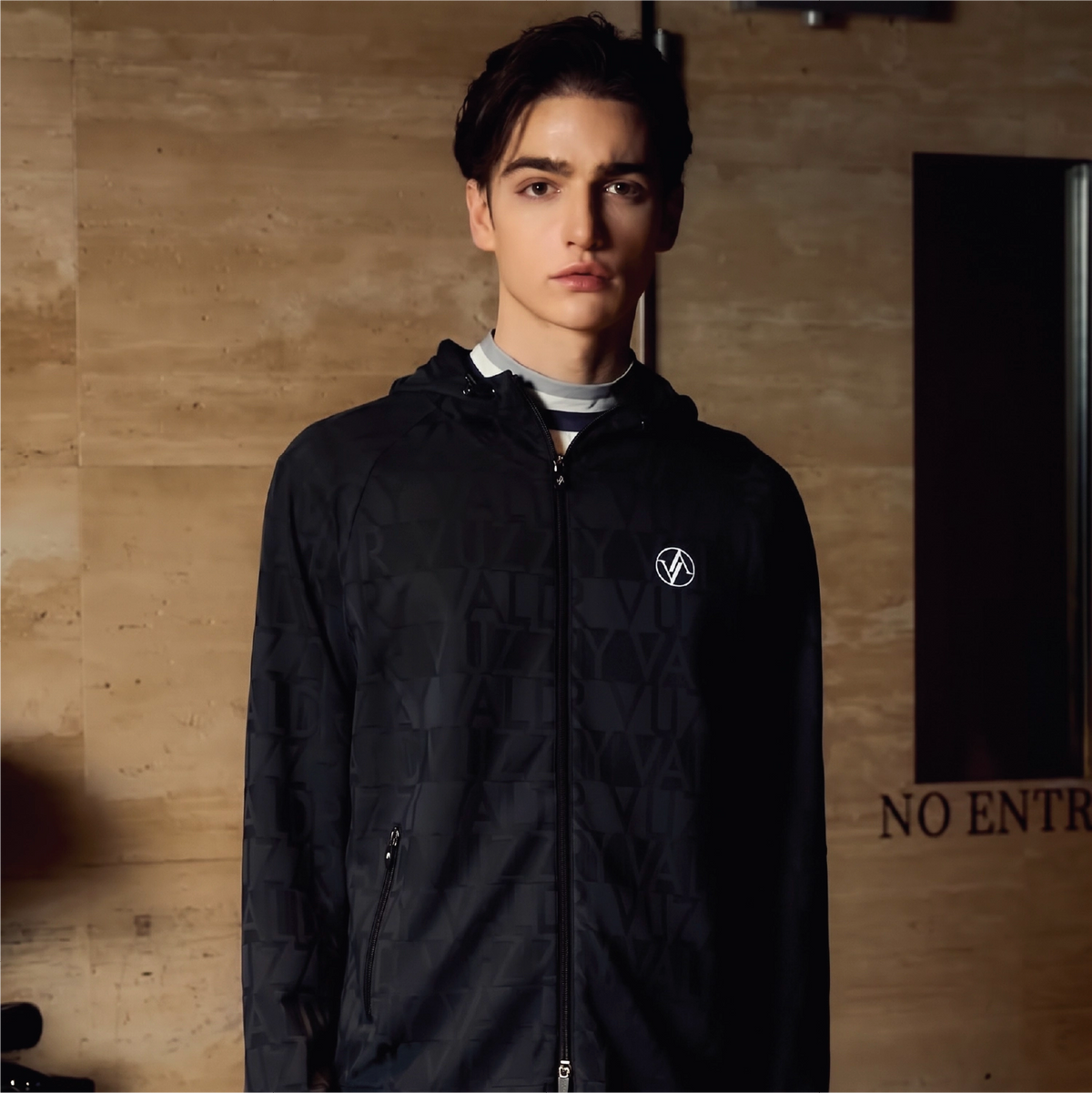 A man wearing a black zip-up hooded jacket with a subtle geometric pattern and a small white logo on the chest, standing against a warm wood-paneled wall.