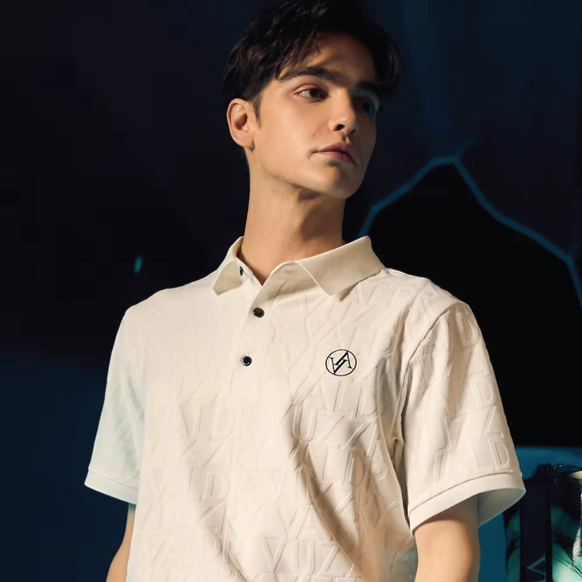 A man wearing a short-sleeve polo shirt with a subtle geometric pattern. The polo is off-white/cream with a small black circular logo on the left chest, set against a dark, moody background.
