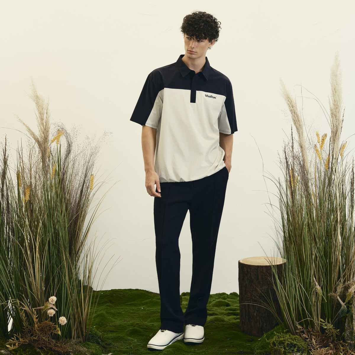 A man stands in a studio wearing a short-sleeve polo shirt with a color-blocked design in navy, cream, and gray, paired with navy pants and white slip-on shoes. The background features a light off-white wall, green moss, and dried pampas grass.