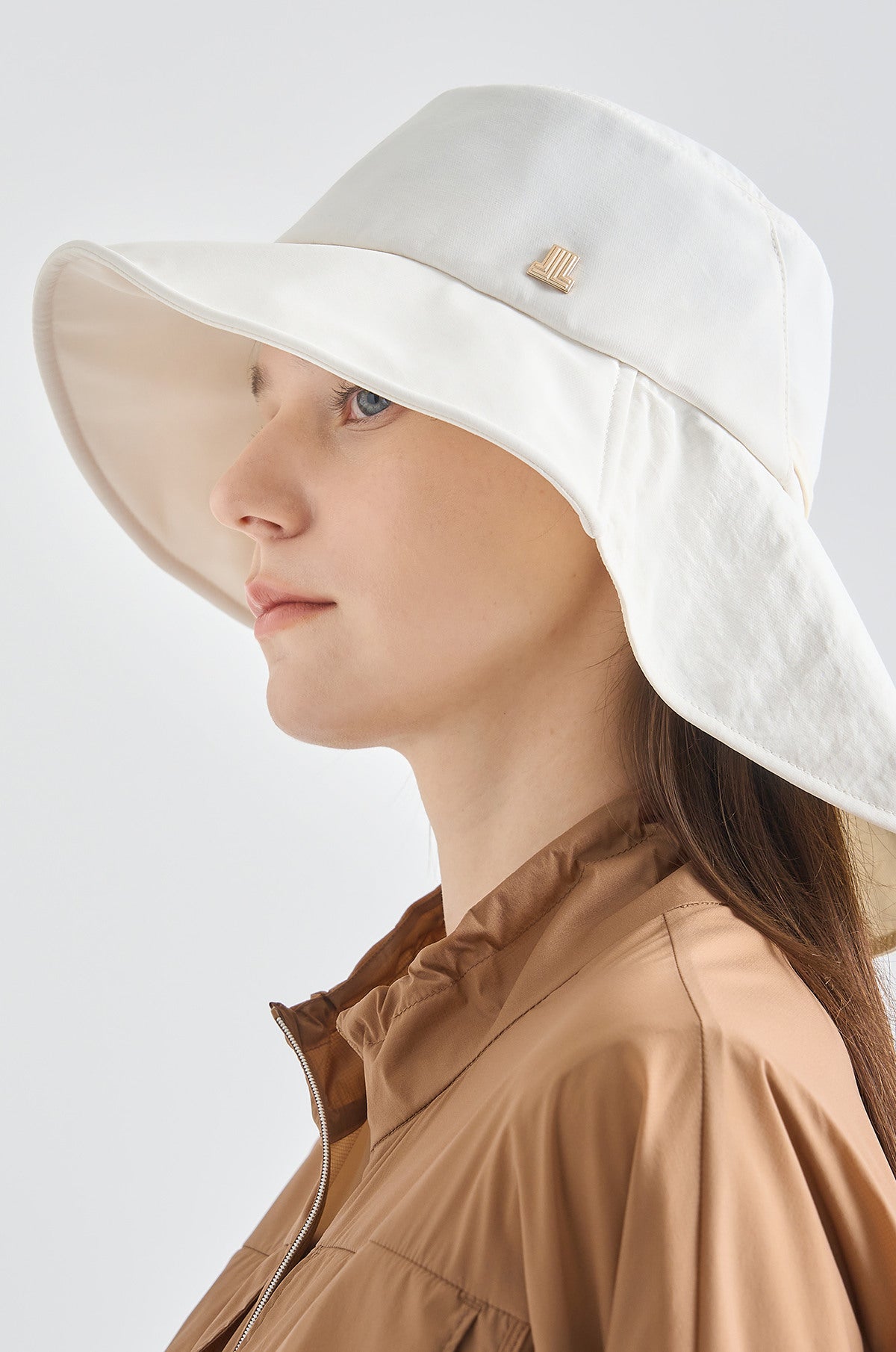 Woman wearing a white wide-brimmed hat and brown jacket on a light gray background