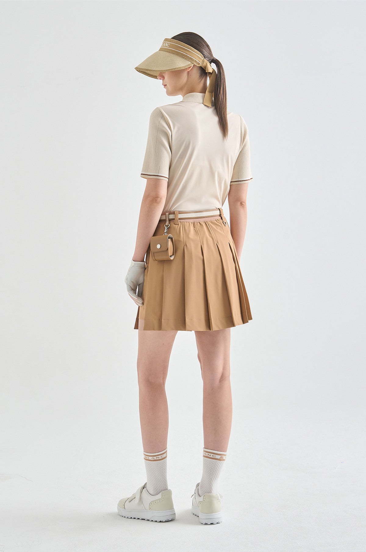 Person wearing a beige visor, white shirt, tan skirt, and white shoes on a light gray background