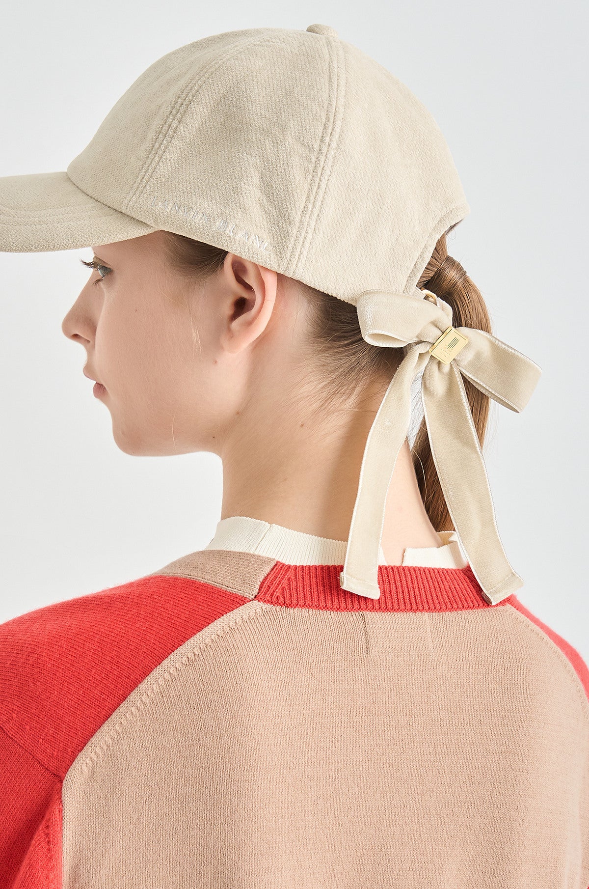 Person wearing a beige cap with a ribbon and a red and beige sweater on a light gray background