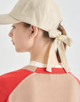 Person wearing a beige cap with a ribbon and a red and beige sweater on a light gray background