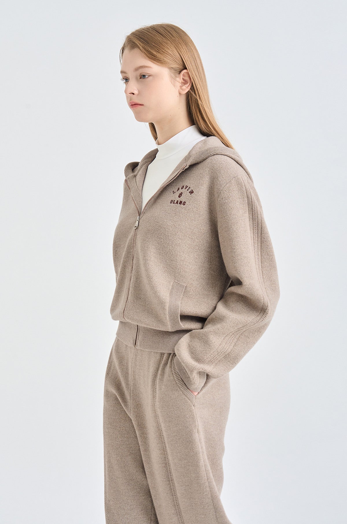 Woman wearing a beige hoodie with a white shirt underneath on a light gray background