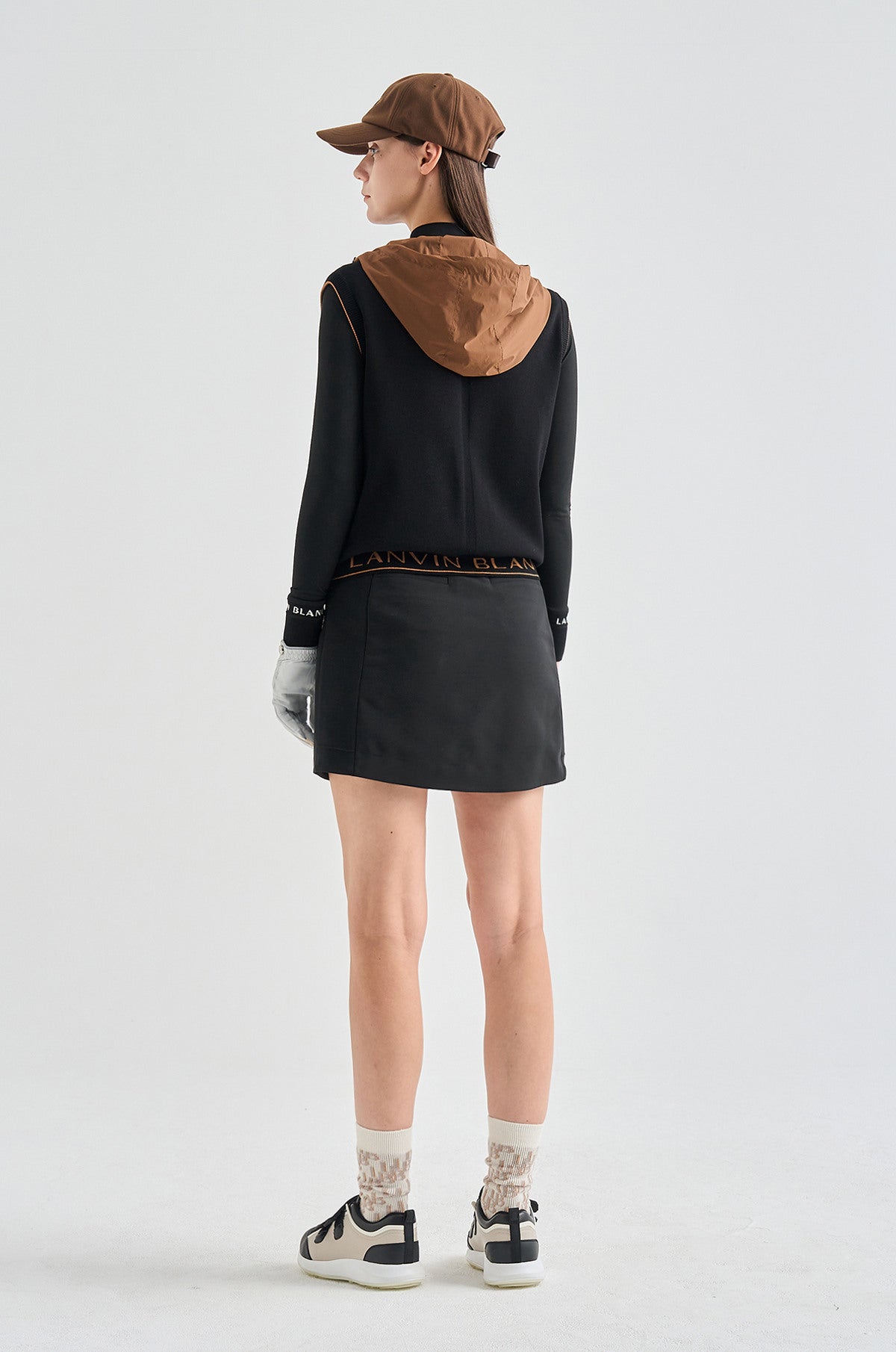 Person wearing a black hoodie with a brown hood, black skirt, and black sneakers on a white background