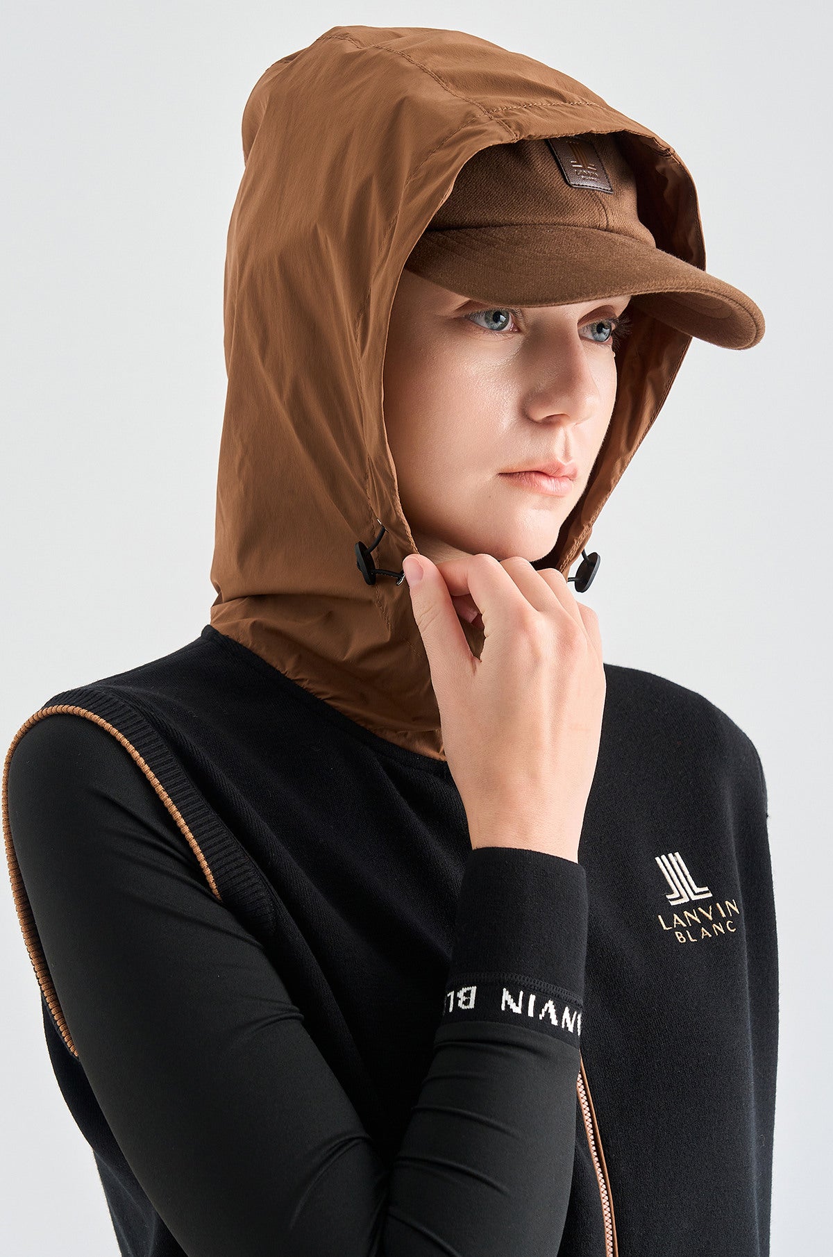 Person wearing a brown hooded jacket and cap with a black long-sleeve shirt featuring a logo.
