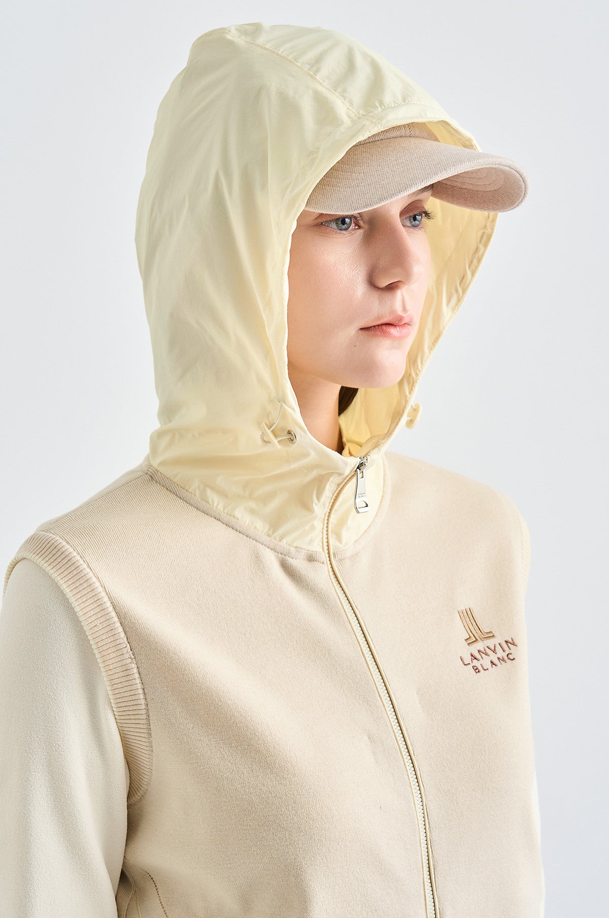 Person wearing a beige hooded jacket with a visible brand logo on a light gray background