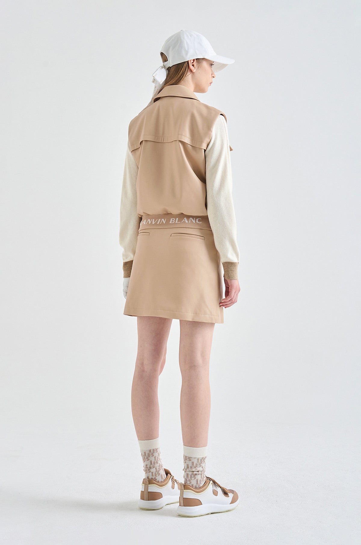 Person wearing a beige jacket and skirt with white top, standing against a white background