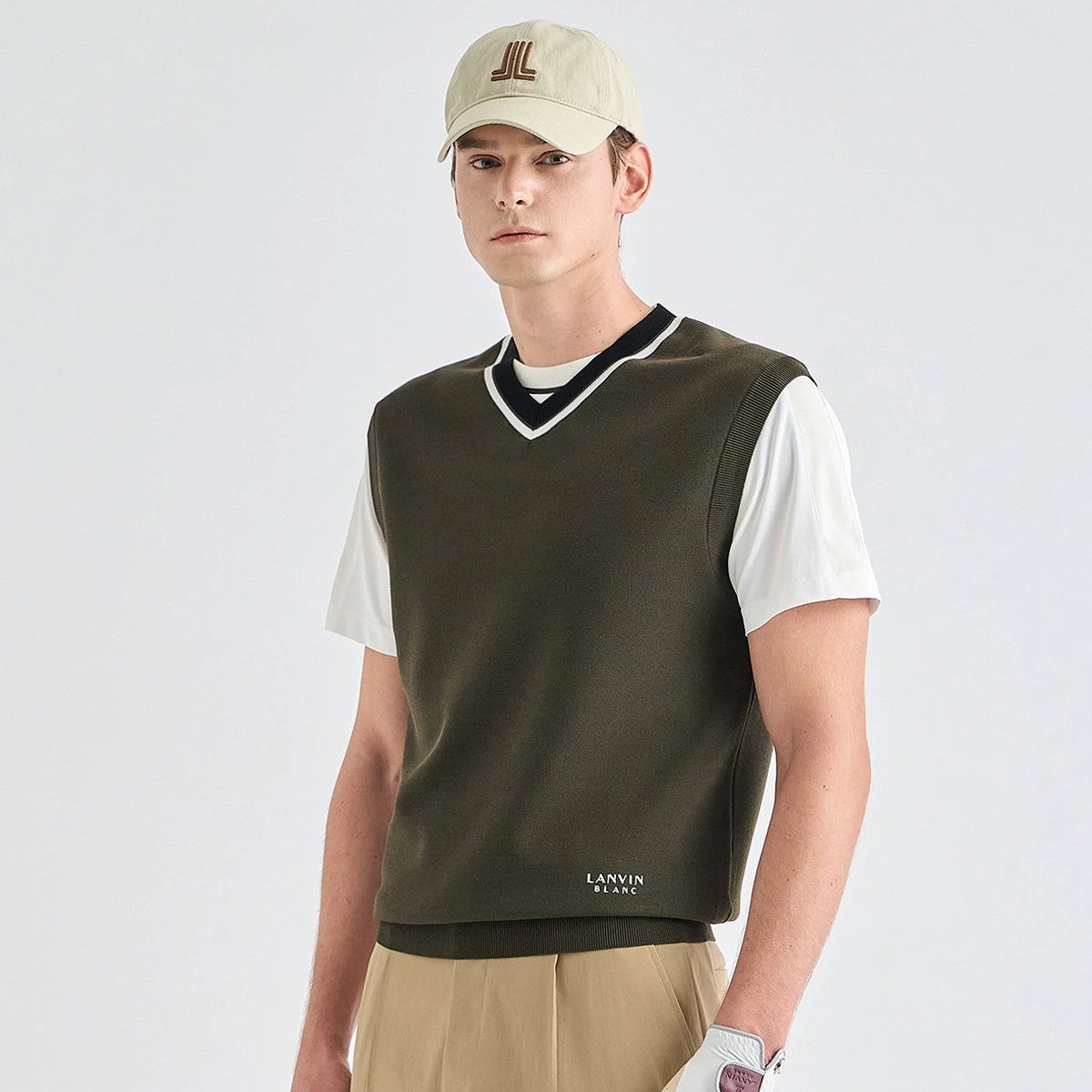 A male model wearing a dark olive green knit sweater vest over a white t-shirt, khaki pants, a beige cap, and a white golf glove, photographed against a light gray studio background.