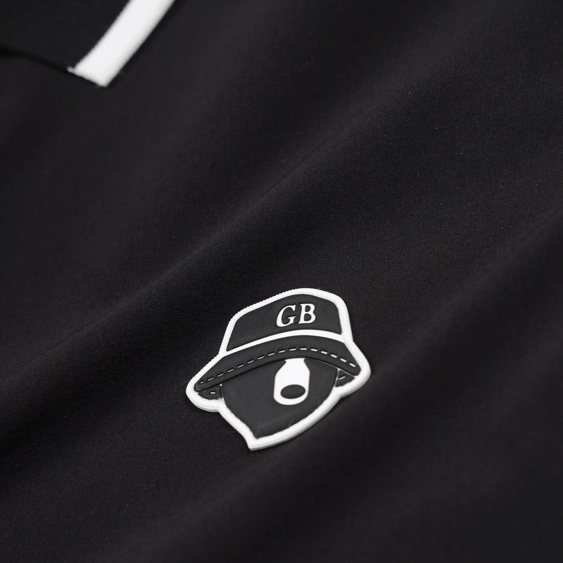 Close-up of a black garment featuring a small embroidered patch with a black-and-white hat design and the letters GB.