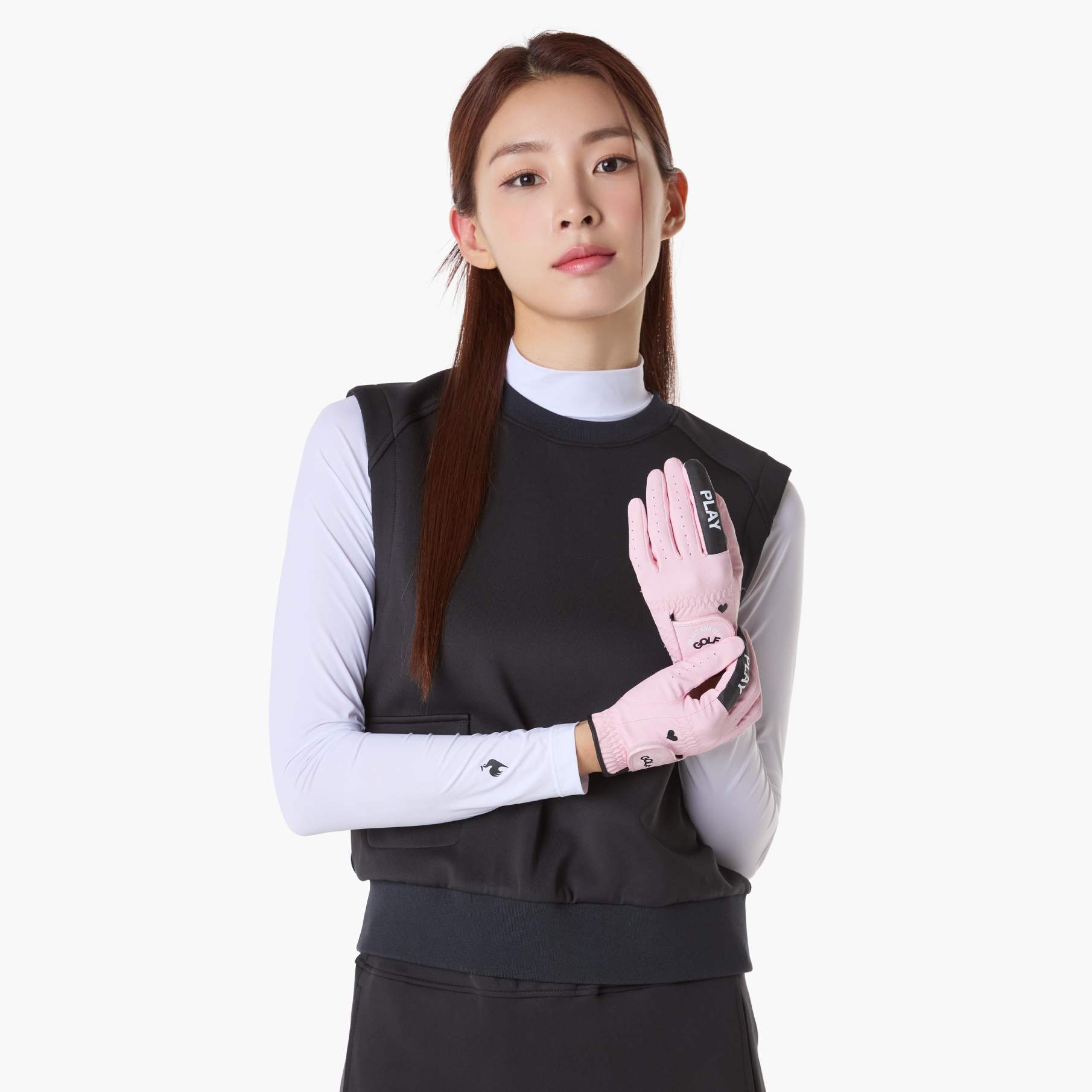 A woman modeling a black athletic sleeveless vest over a white long-sleeve top, wearing light pink sports gloves with black branding, against a plain light gray background.