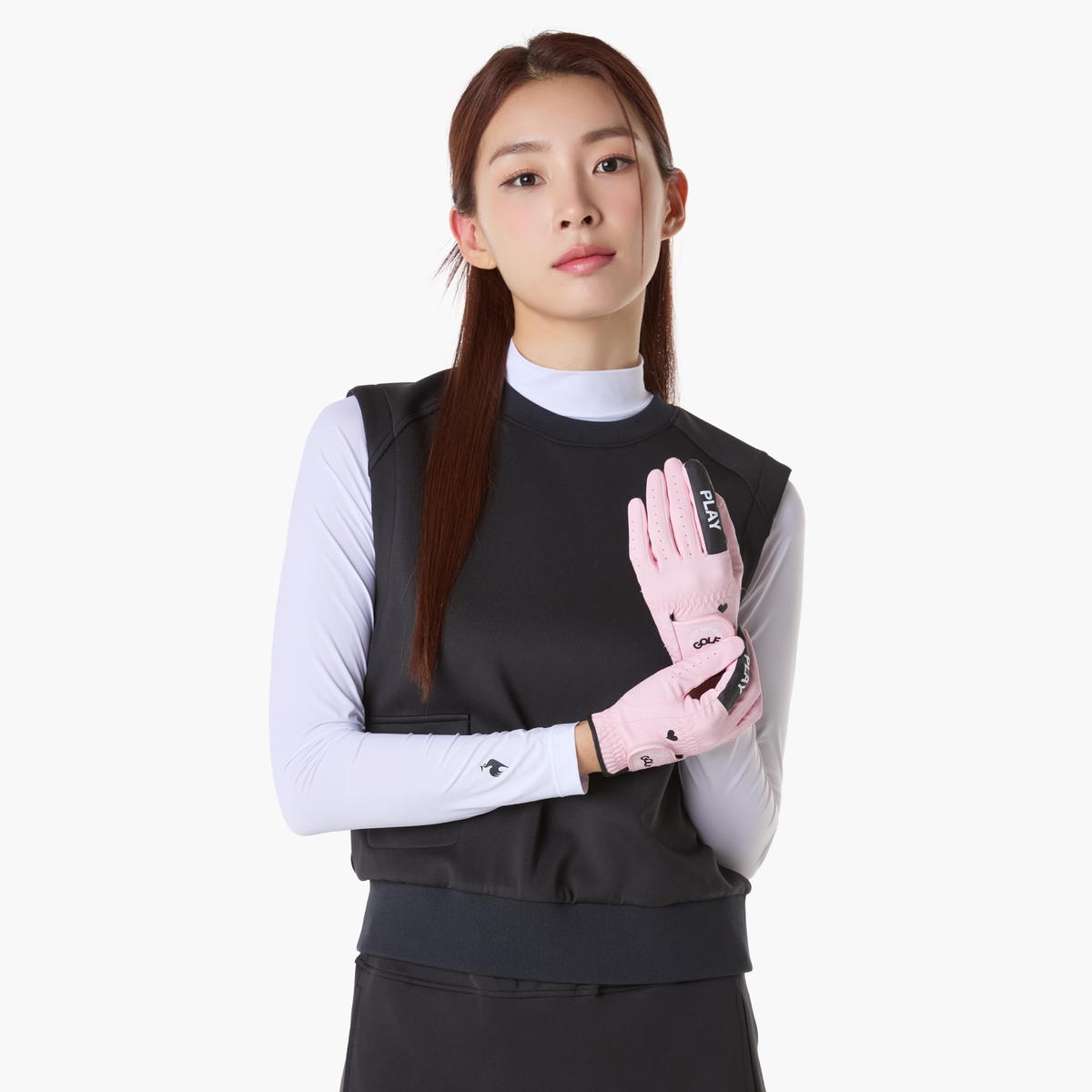 A woman modeling a black athletic sleeveless vest over a white long-sleeve top, wearing light pink sports gloves with black branding, against a plain light gray background.