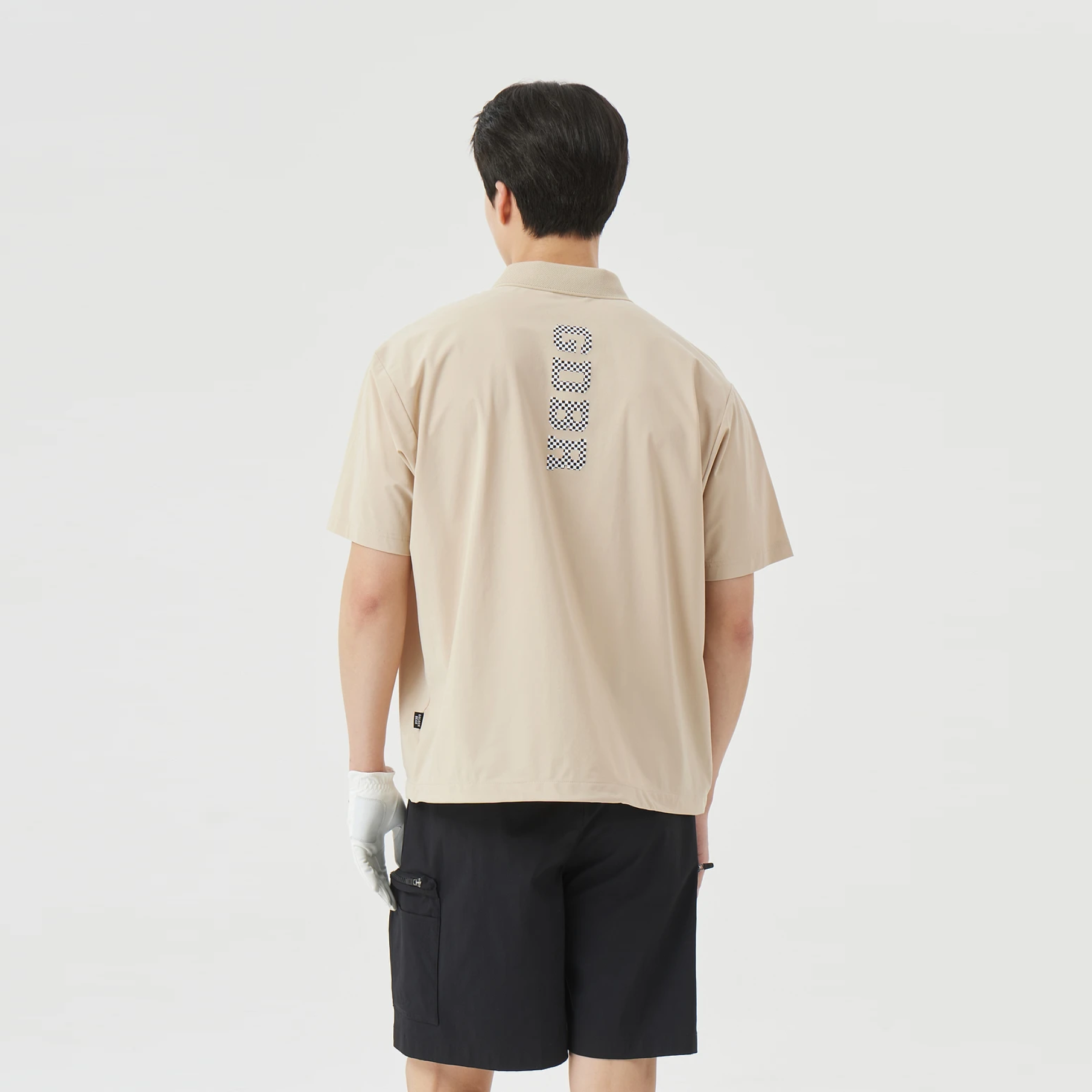 A man viewed from behind wearing a beige short-sleeve polo shirt with a vertical patterned logo on the back and black shorts, against a plain light gray studio background.