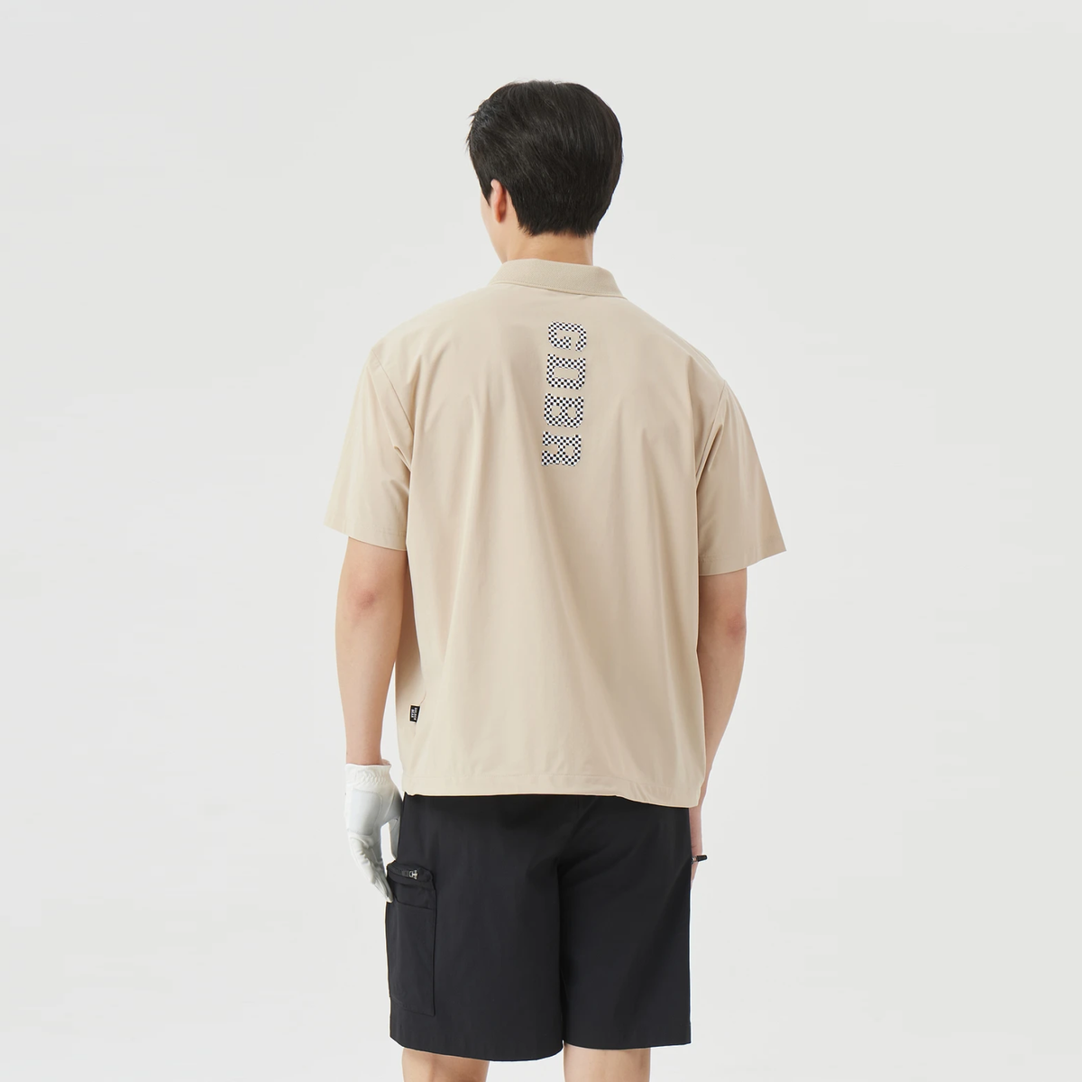 A man viewed from behind wearing a beige short-sleeve polo shirt with a vertical patterned logo on the back and black shorts, against a plain light gray studio background.
