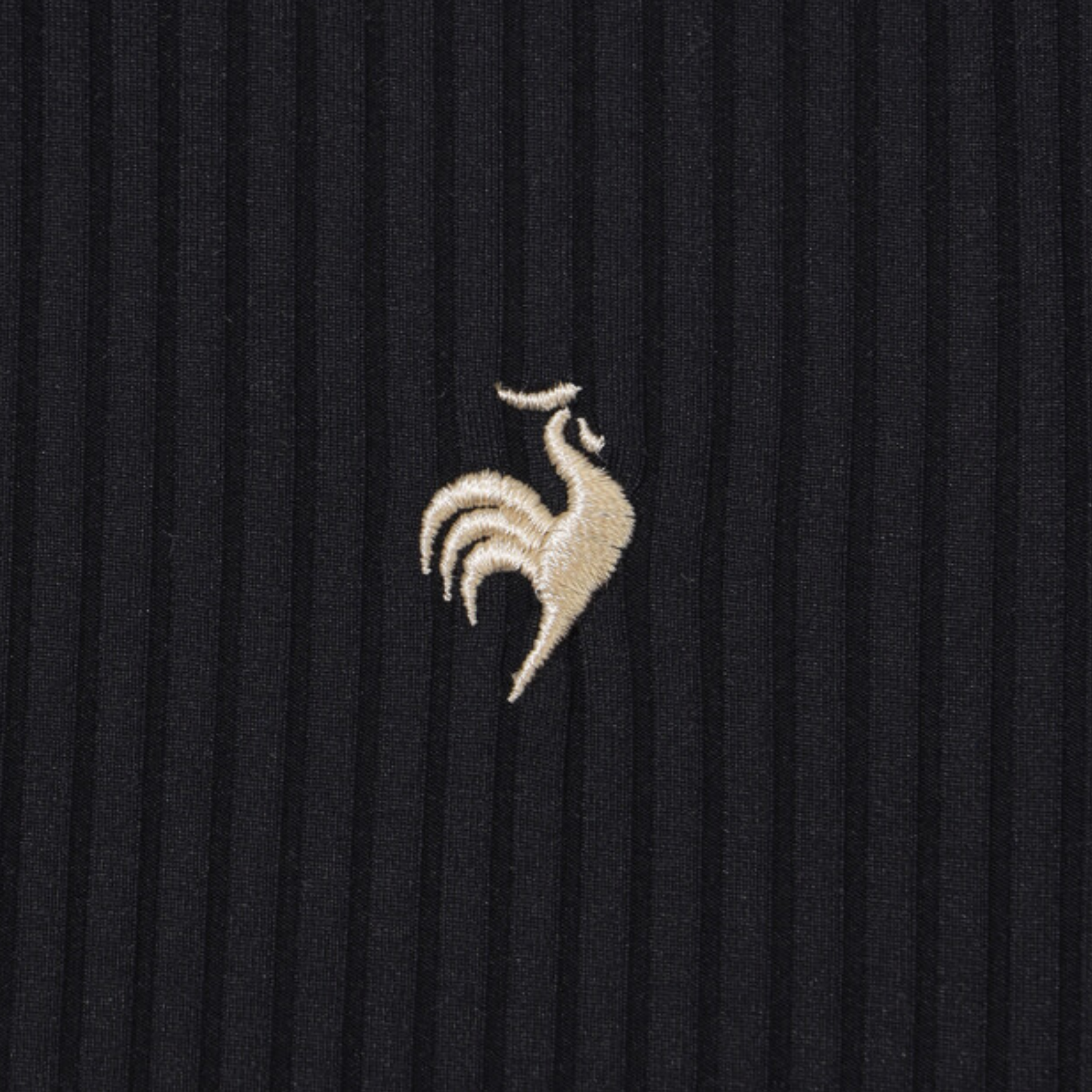 Close-up of a ribbed black knit fabric with a small embroidered rooster logo in a light beige or cream tone at the center.