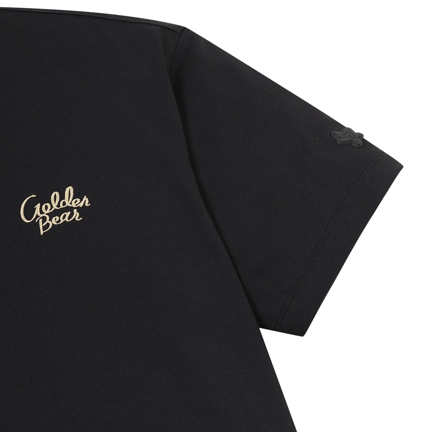 Close-up of a black short-sleeve T-shirt showing the left chest area with a gold 'Golden Bear' script logo and a small tonal black logo on the sleeve, against a white background.