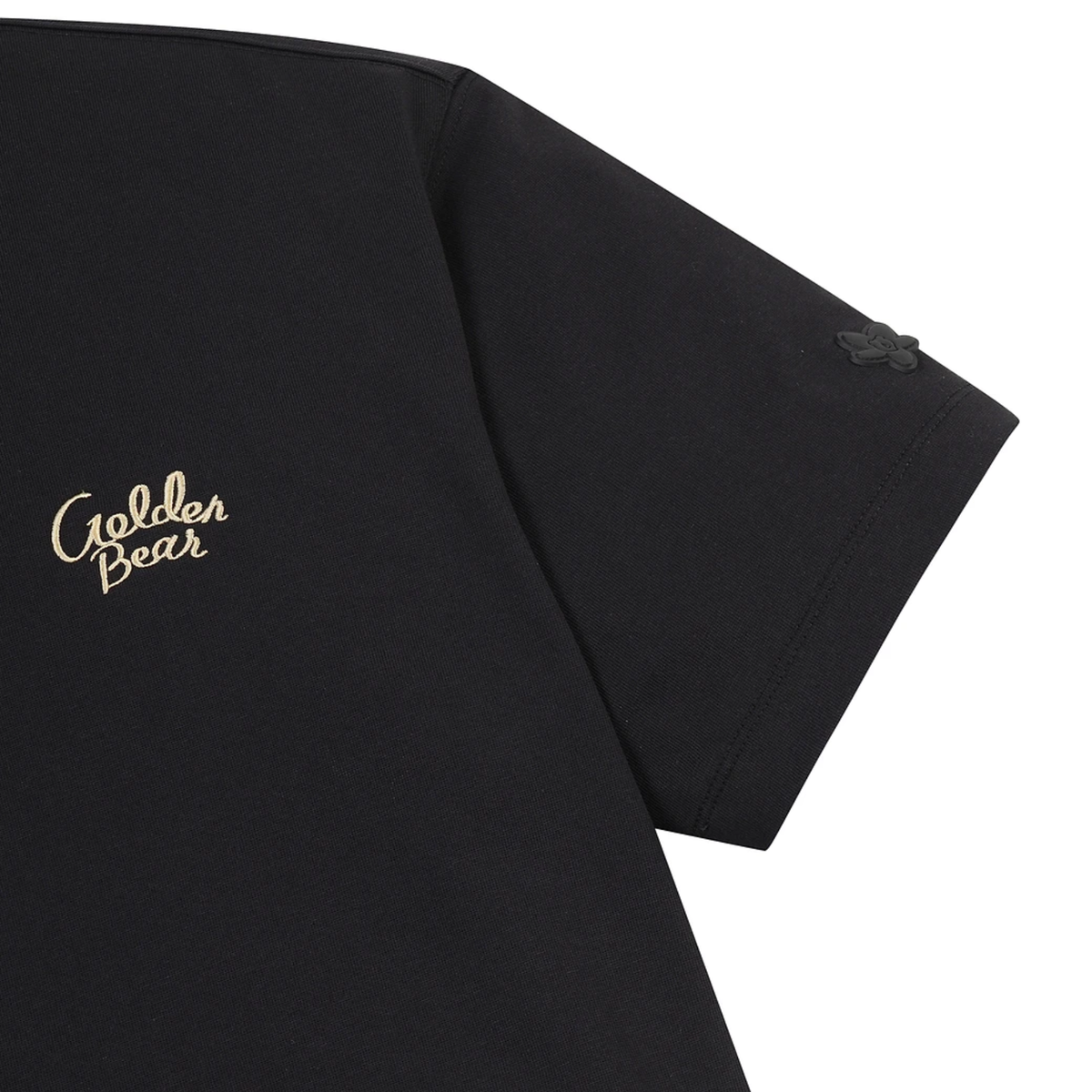 Close-up of a black short-sleeve T-shirt showing the left chest area with a gold 'Golden Bear' script logo and a small tonal black logo on the sleeve, against a white background.