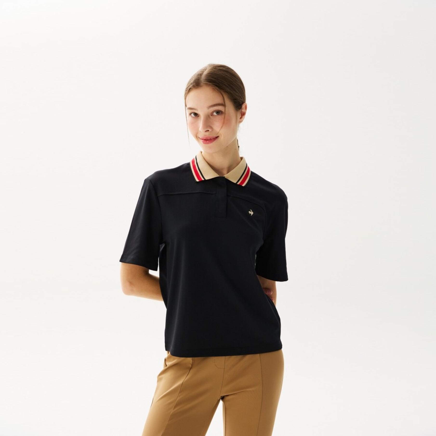 A woman modeling a black short-sleeve polo shirt with a beige, red, and white striped collar, paired with tan trousers, against a plain white studio background.