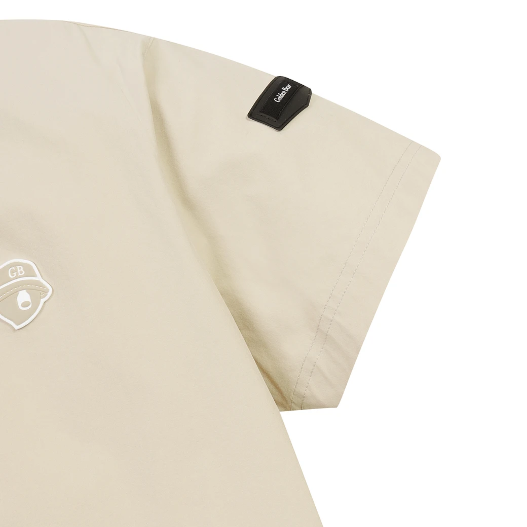 Close-up of a light beige short-sleeve garment, showing part of the sleeve with a black brand tab and a small white chest patch on a plain white background.