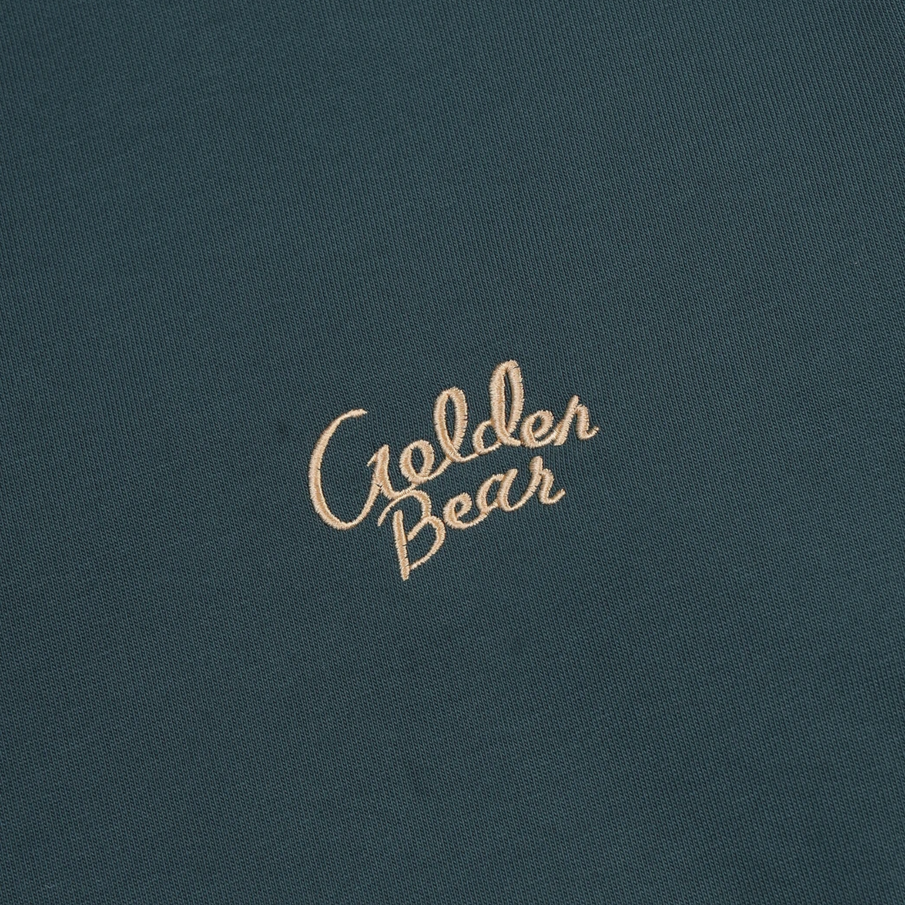 Close-up of a dark teal fabric surface with the words 'Golden Bear' embroidered in a light tan script near the center.