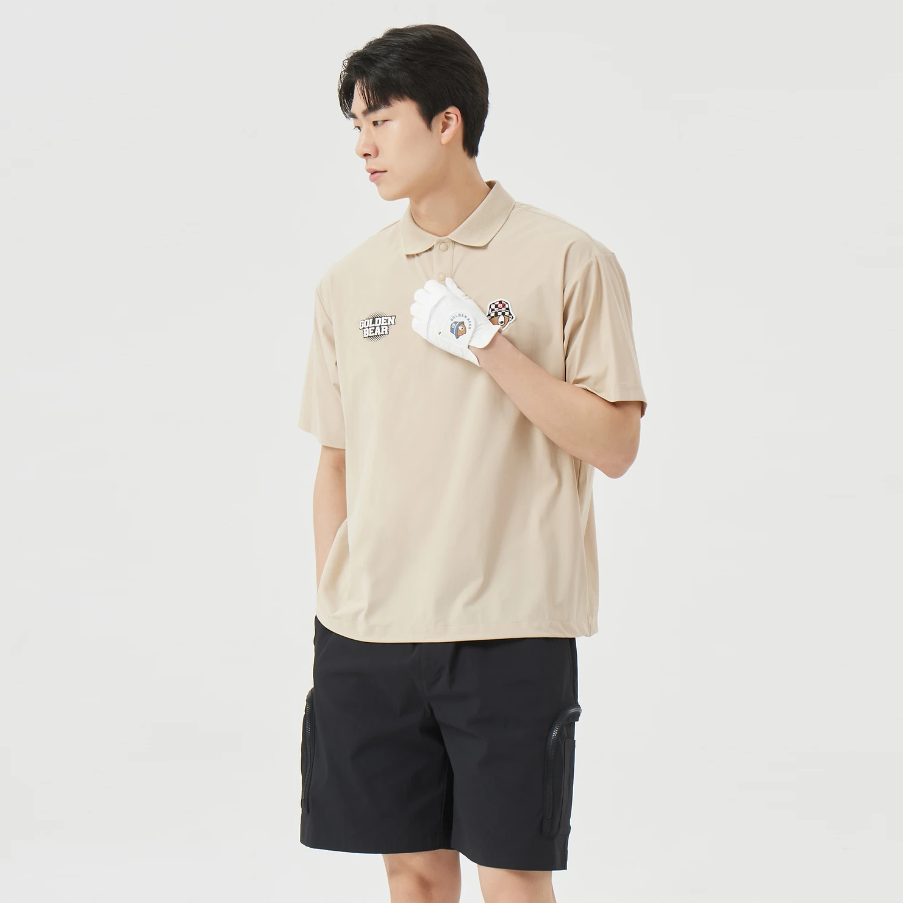 A man wearing a beige short-sleeve polo shirt with a small graphic on the left chest, black cargo shorts, and a white golf glove, standing against a light gray studio background.