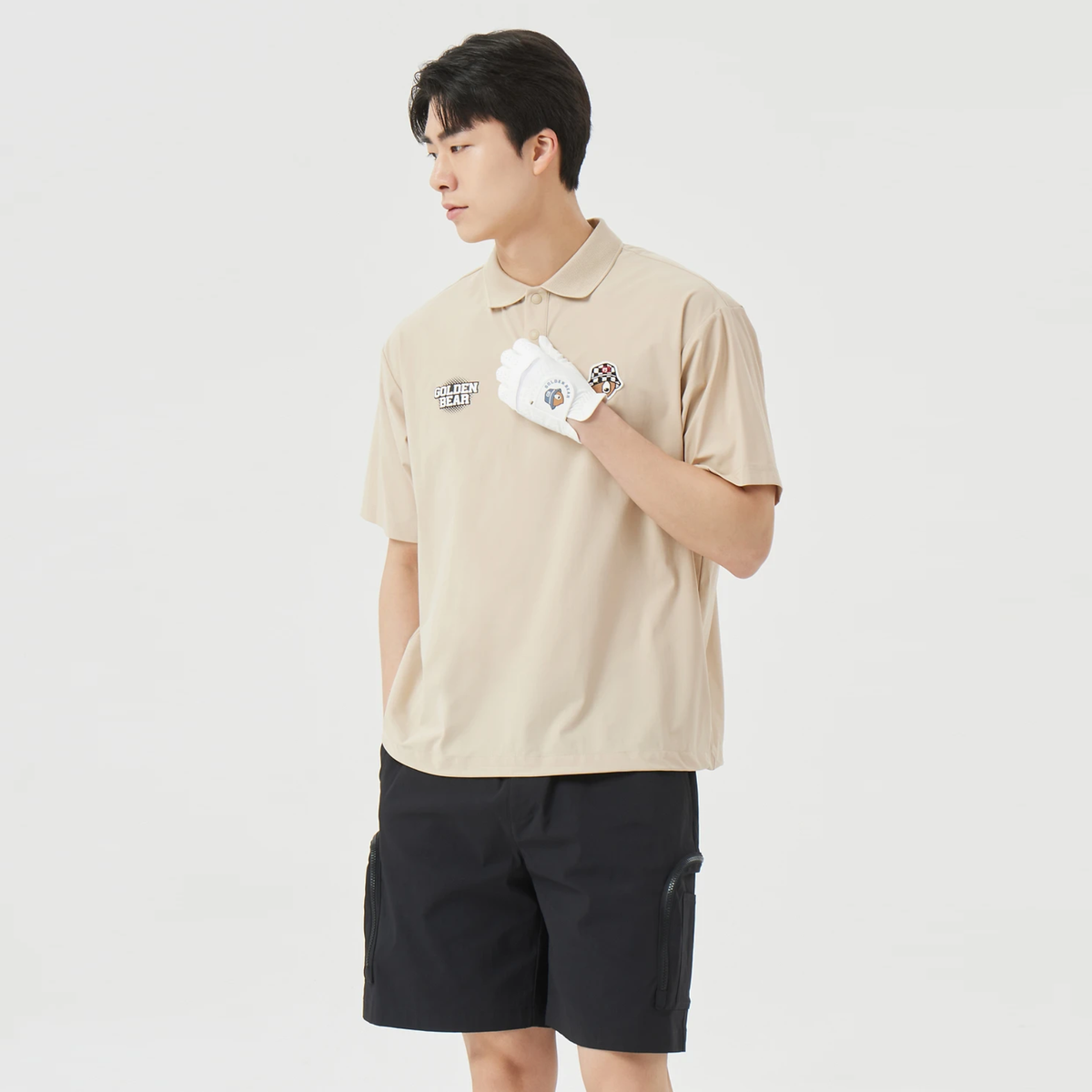 A man wearing a beige short-sleeve polo shirt with a small graphic on the left chest, black cargo shorts, and a white golf glove, standing against a light gray studio background.