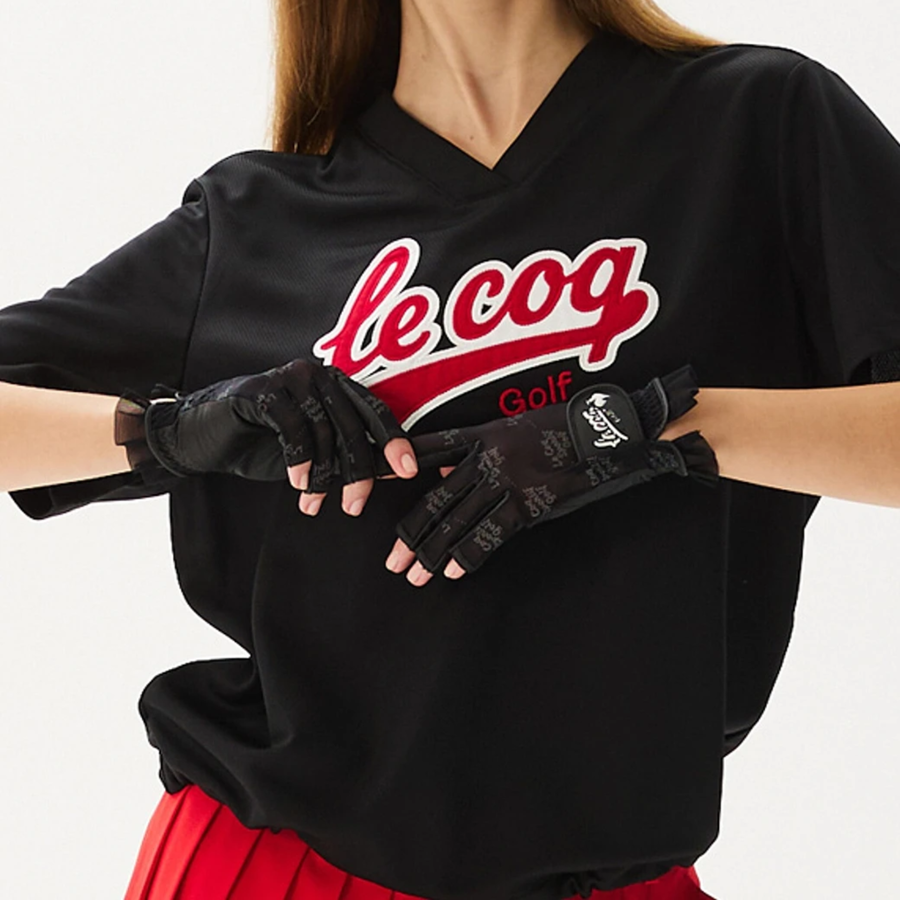 A person wearing a black V-neck T-shirt with a red and white 'le coq Golf' logo and black fingerless golf gloves, shown against a plain light background.