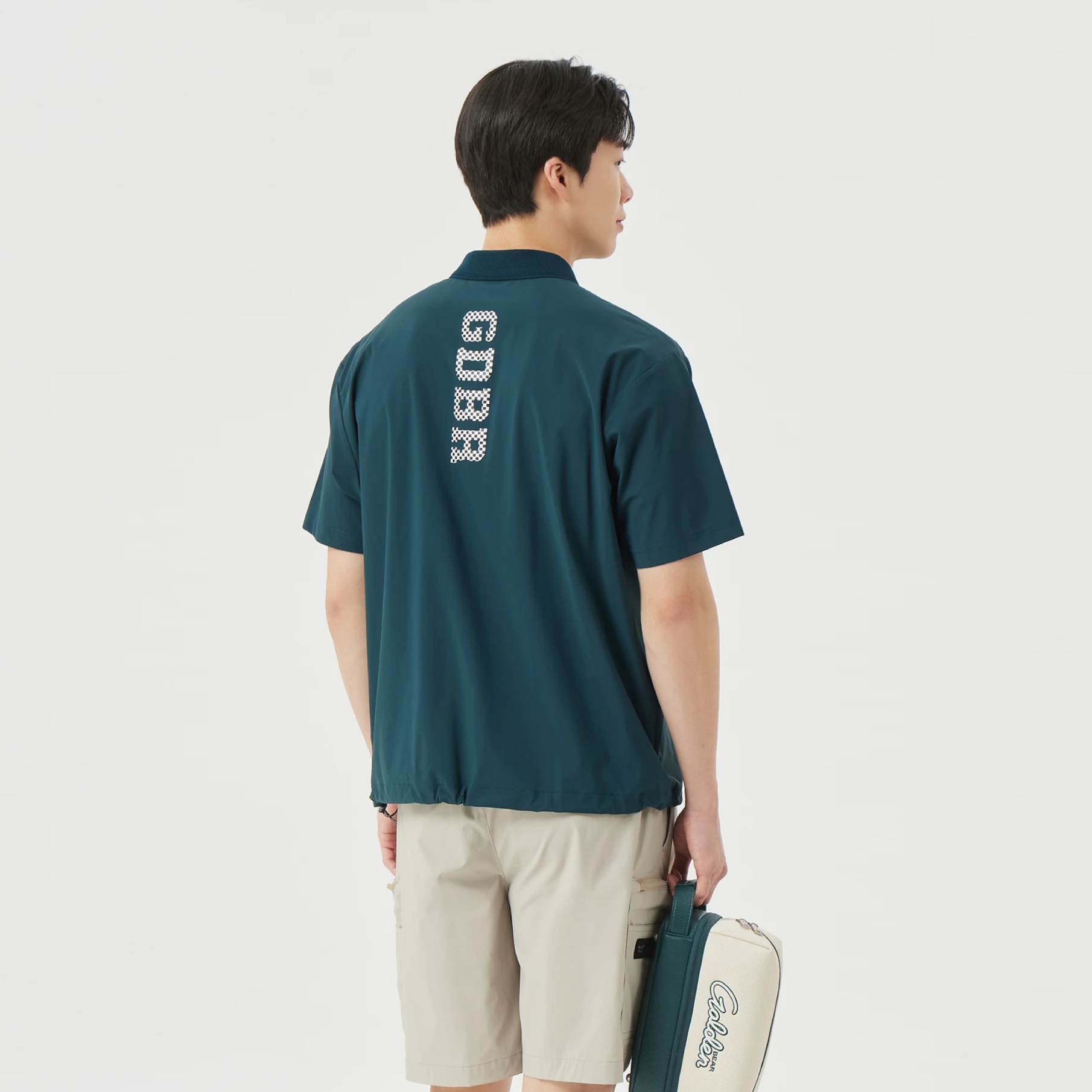 A man is shown from the back wearing a dark teal short-sleeve collared shirt with 'GUBR' printed vertically in white on the back, paired with beige shorts and holding a matching teal and white bag against a light gray studio background.