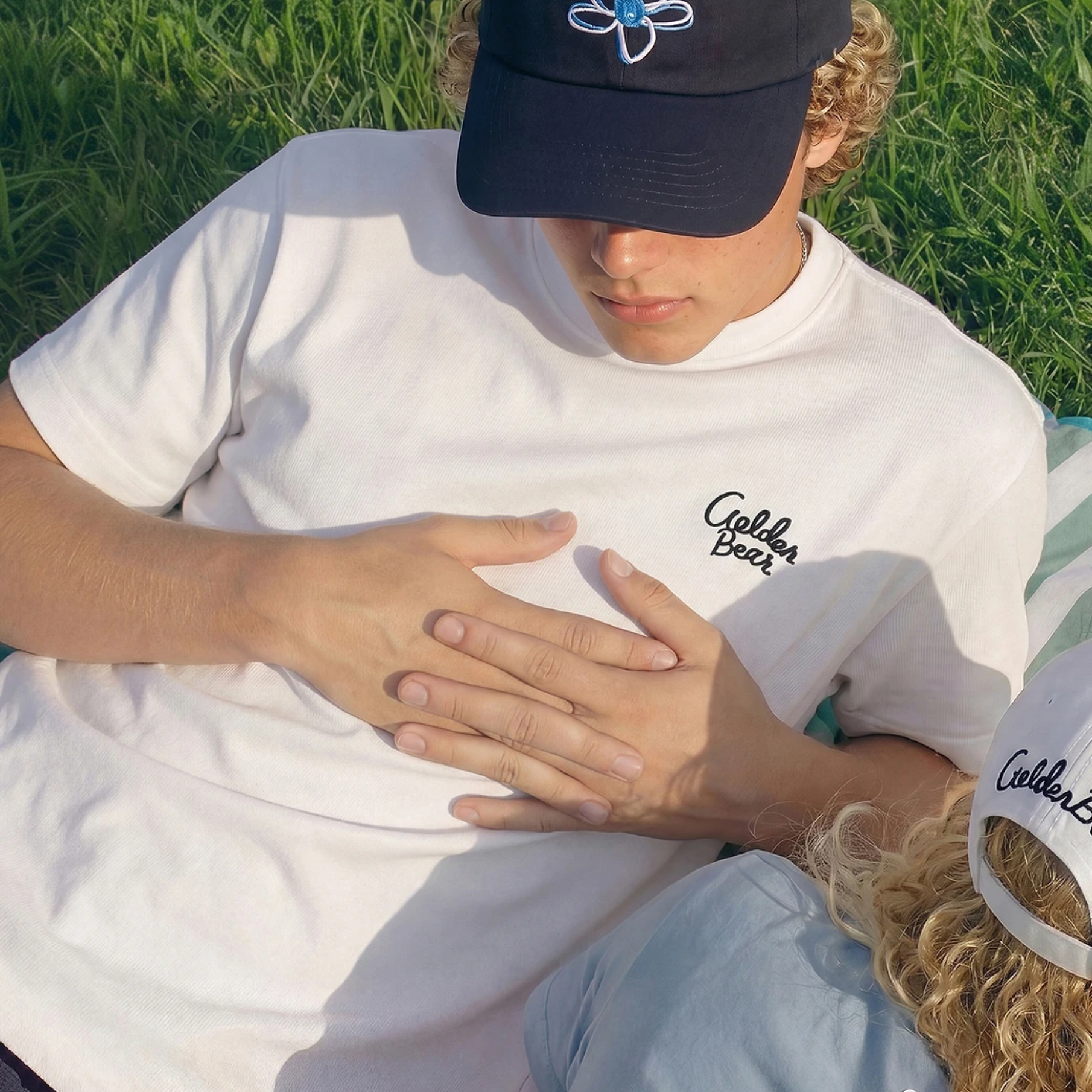 A person outdoors on grass wearing a white T-shirt with a small black script logo on the chest and a black baseball cap with a blue flower logo. Another person is partially visible in the foreground wearing a white cap with the same script.