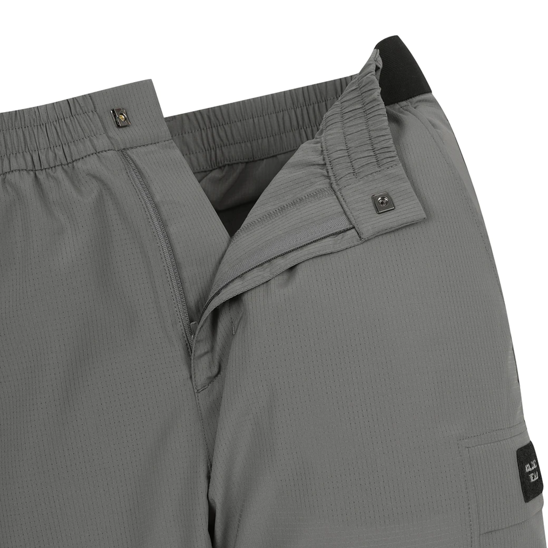 Close-up of gray technical outdoor pants showing the elastic waistband, reinforced pocket seam, and a small black brand patch on the lower leg.