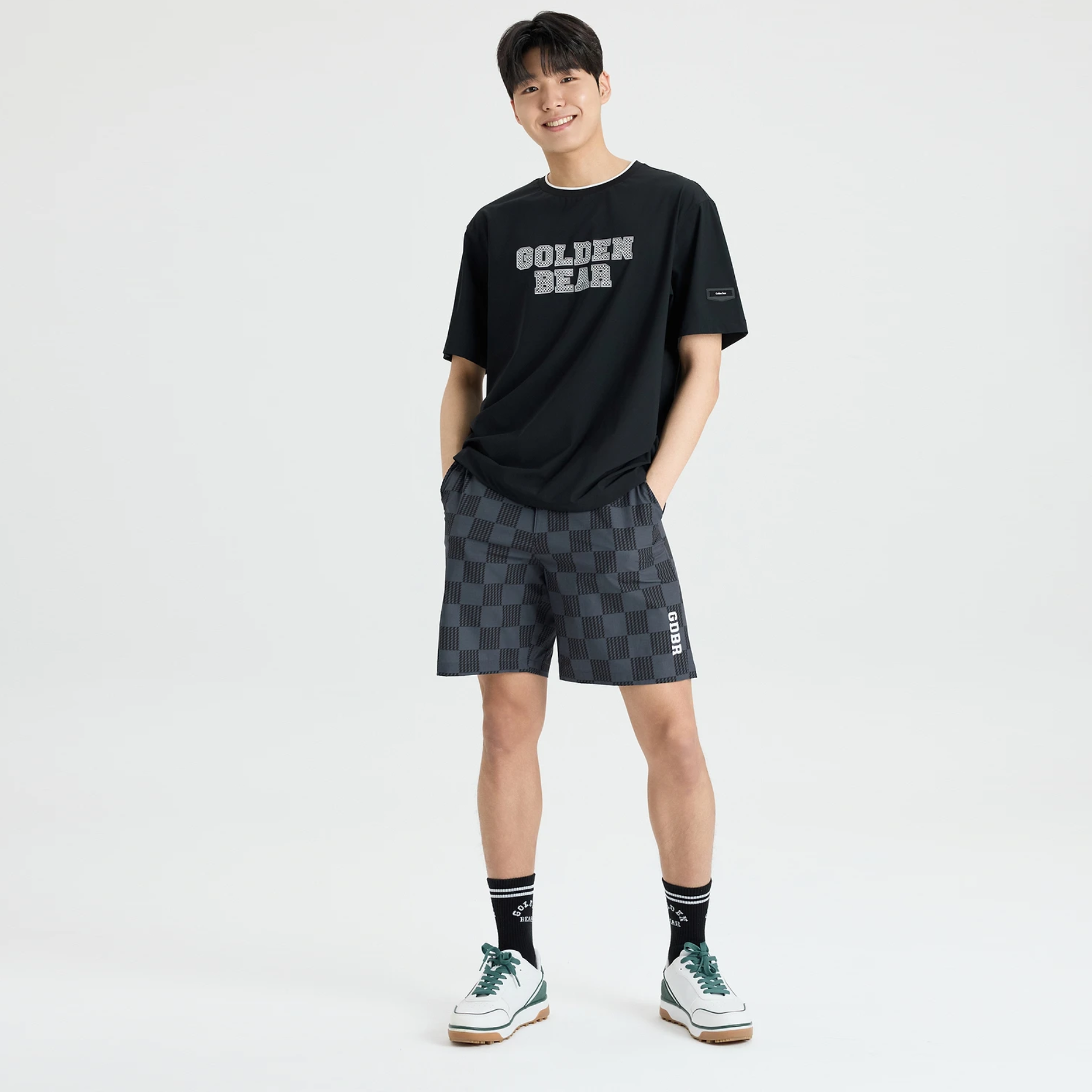 A man modeling a black t-shirt with 'GOLDEN BEAR' text and dark gray checkered shorts, standing against a light gray studio background.