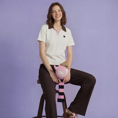 A woman sits on a stool wearing a cream short-sleeve polo shirt with a brown collar and a small black embroidered logo, paired with dark brown trousers. She holds a pink plush toy against her lap.