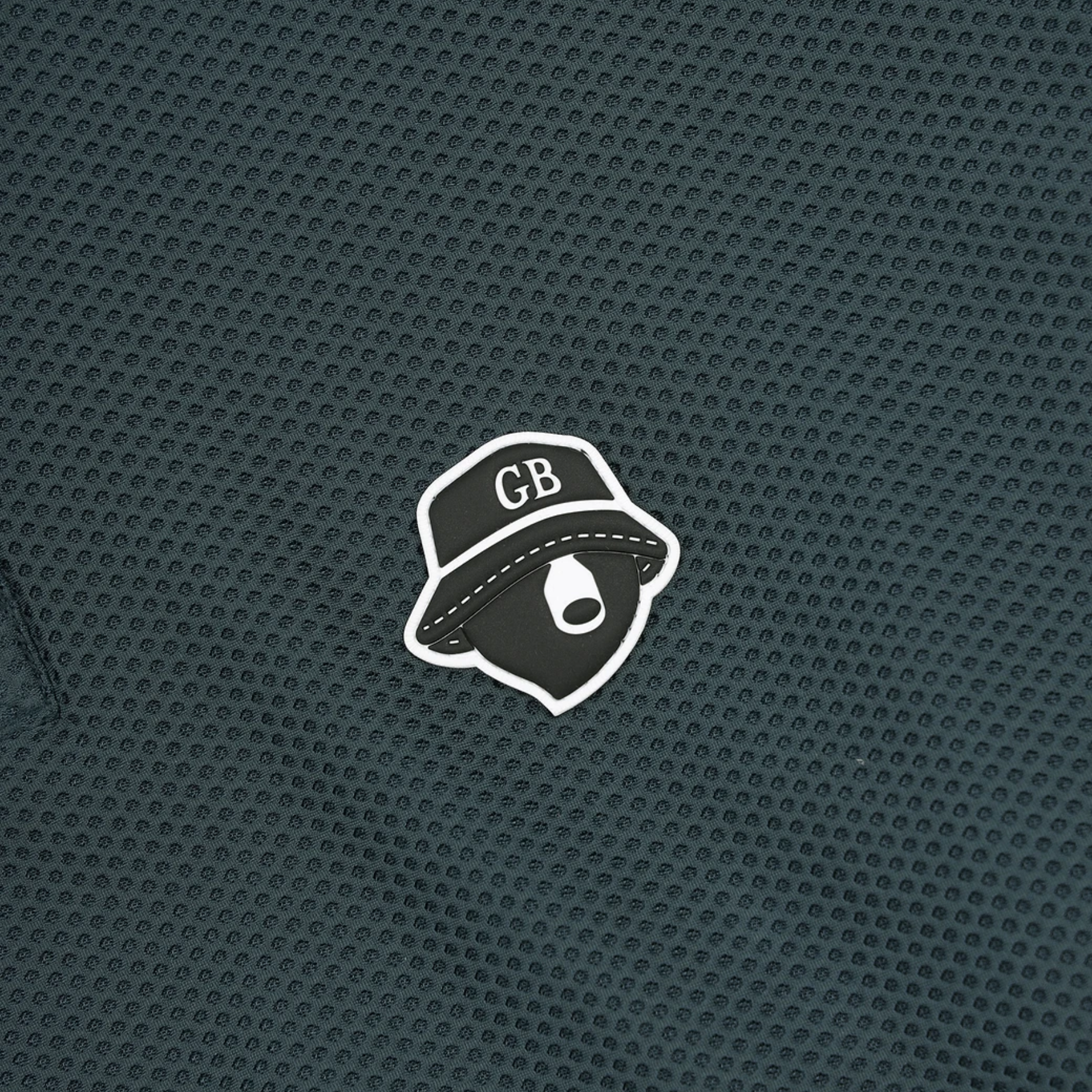 A close-up of a dark teal textured mesh fabric featuring a small white and black embroidered hat patch with the letters 'GB' and a stylized eye logo.
