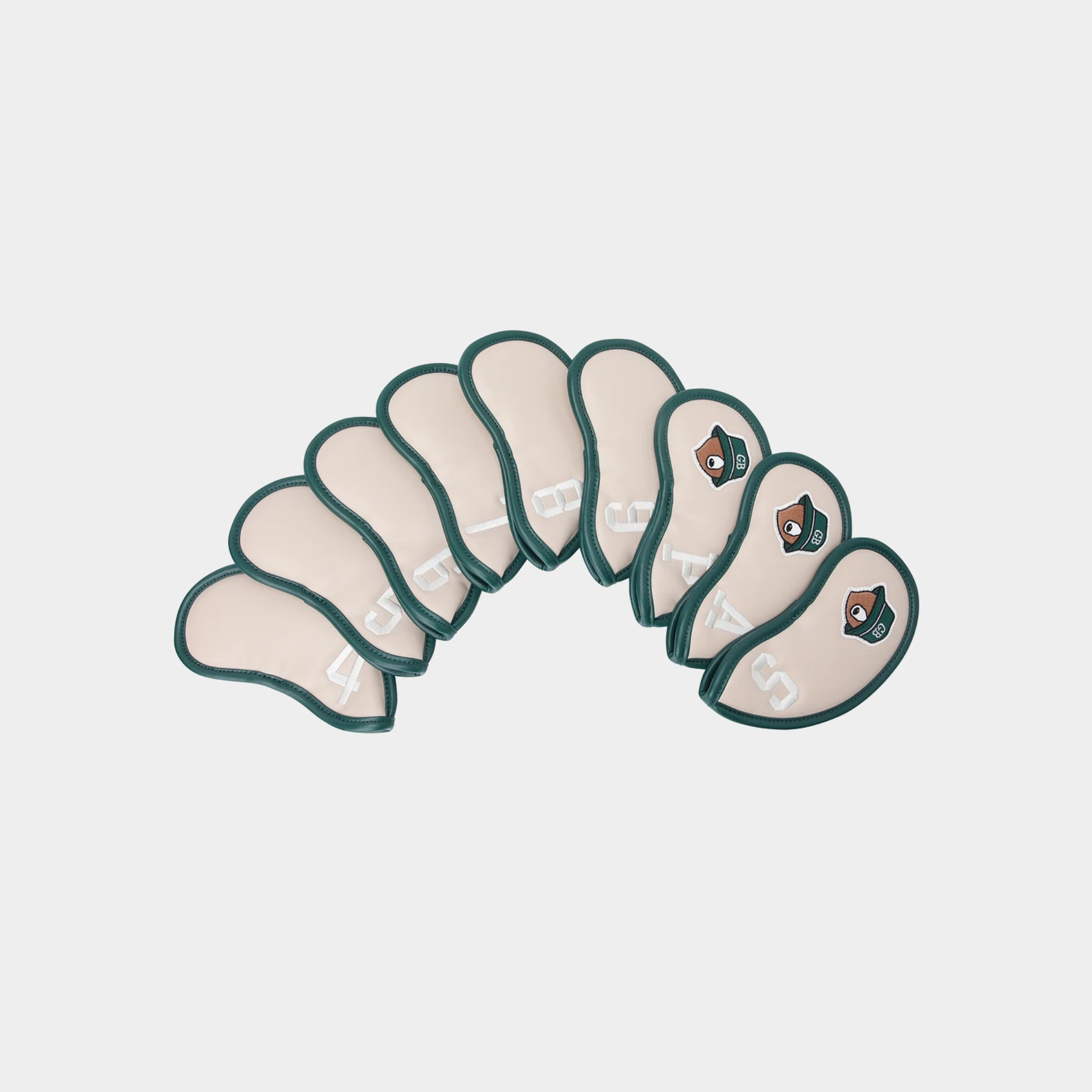 A set of nine golf club headcovers arranged in a semi-circle on a light gray background. The covers are a warm cream color with a dark teal-green stitched border and a small embroidered geometric logo near the base of each cover.