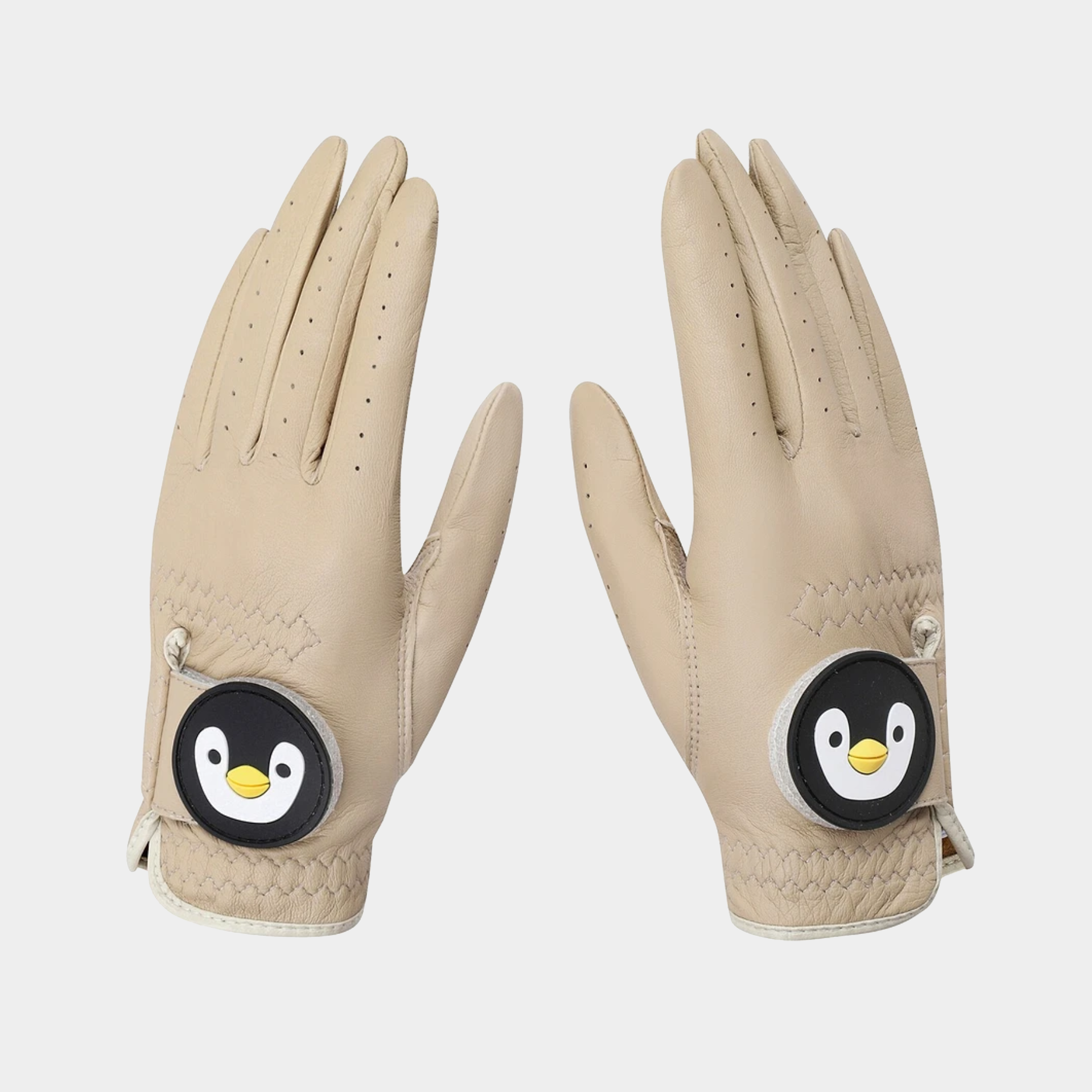 A pair of beige leather golf gloves with a circular penguin patch on the back of the hand, shown against a light gray background.
