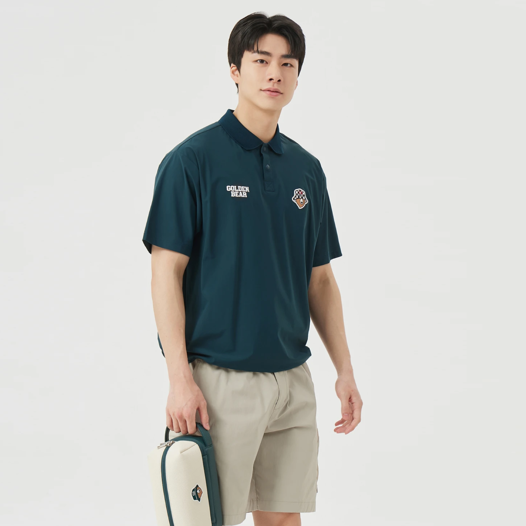 A man wearing a dark teal polo shirt with embroidered logos on the chest, paired with beige shorts, holding a matching dark teal and cream bag against a light gray studio background.