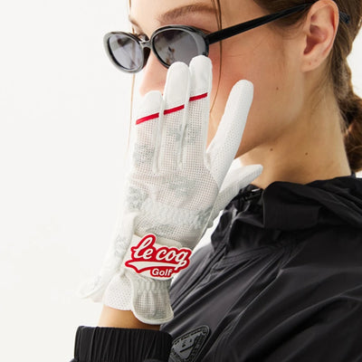 A close-up lifestyle shot of a model wearing a white Le Coq Golf glove with red accents and sunglasses, paired with a black jacket against a clean white background.